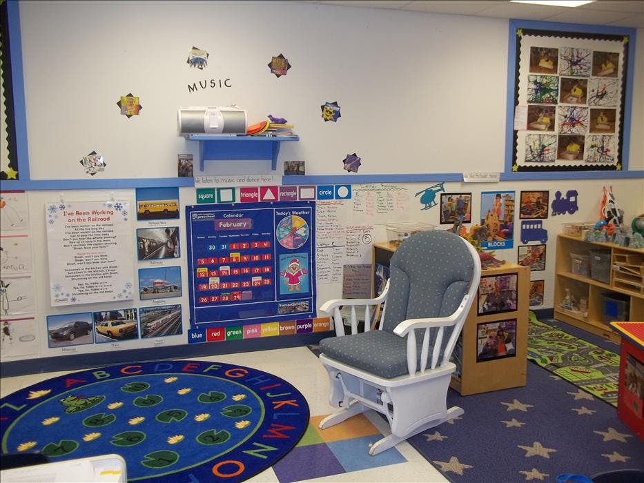 A classroom with a calendar chart, a rocking chair, and a frog rug on the floor