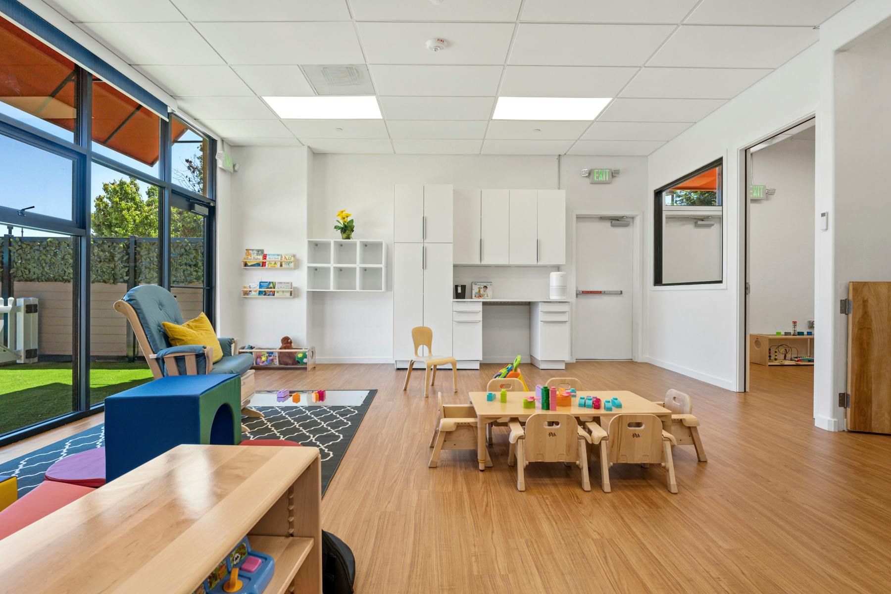 Empty classroom with small tables and chairs, large windows, and a reading nook