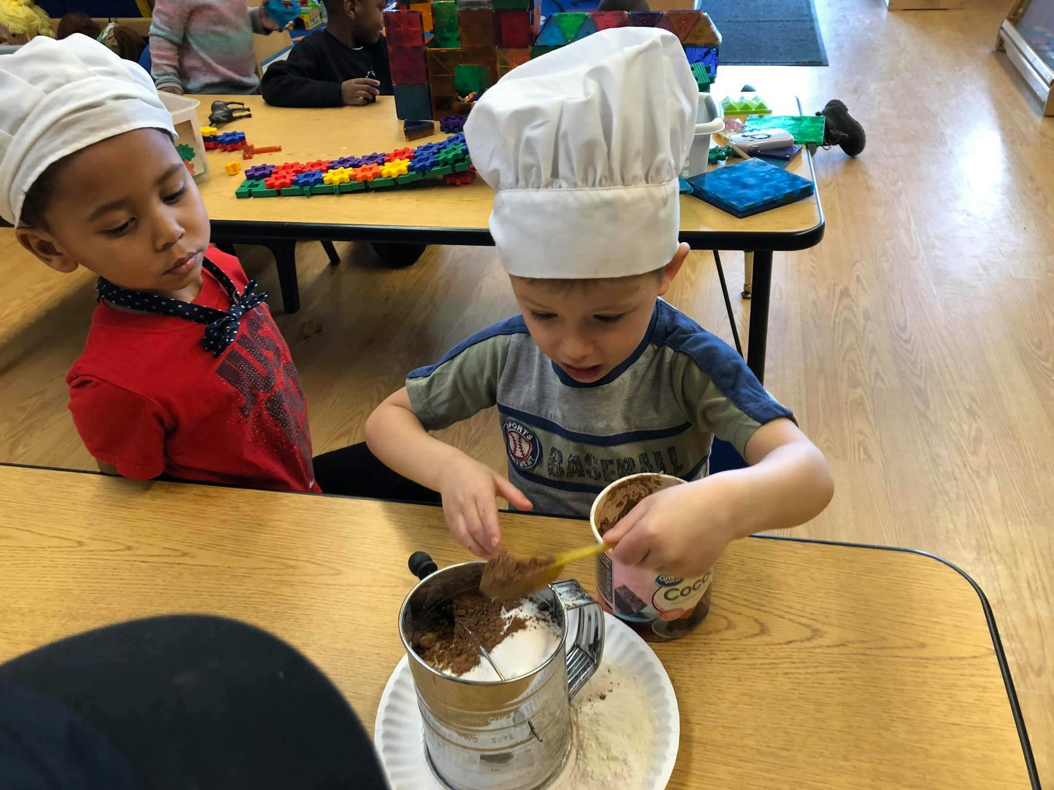 Two children in chef hats at a table, one sifting ingredients