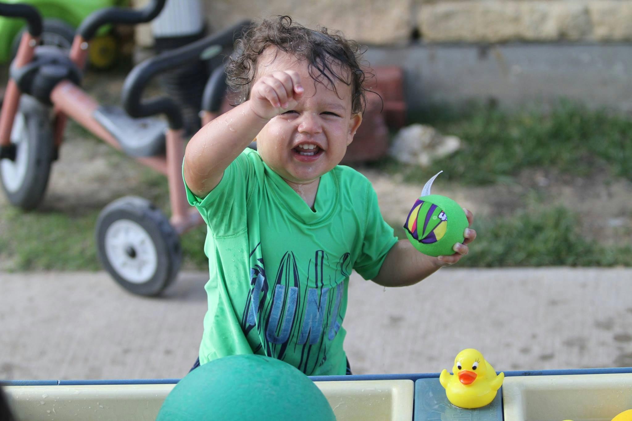 Child in green shirt holding a green toy, with a yellow rubber duck nearby