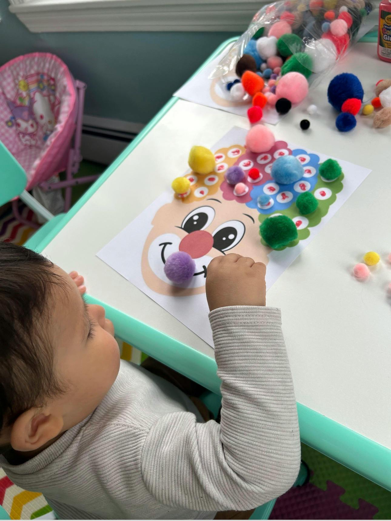Child crafting with colorful pom poms on a clown face drawing