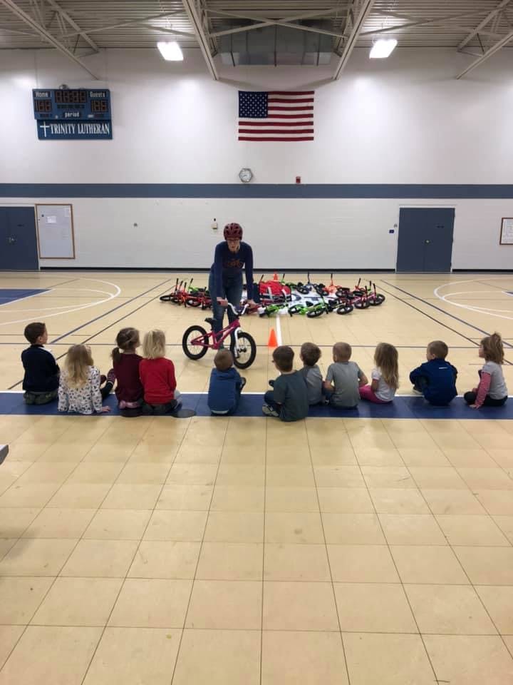 Person on a small bicycle in a gym with children sitting on the floor watching