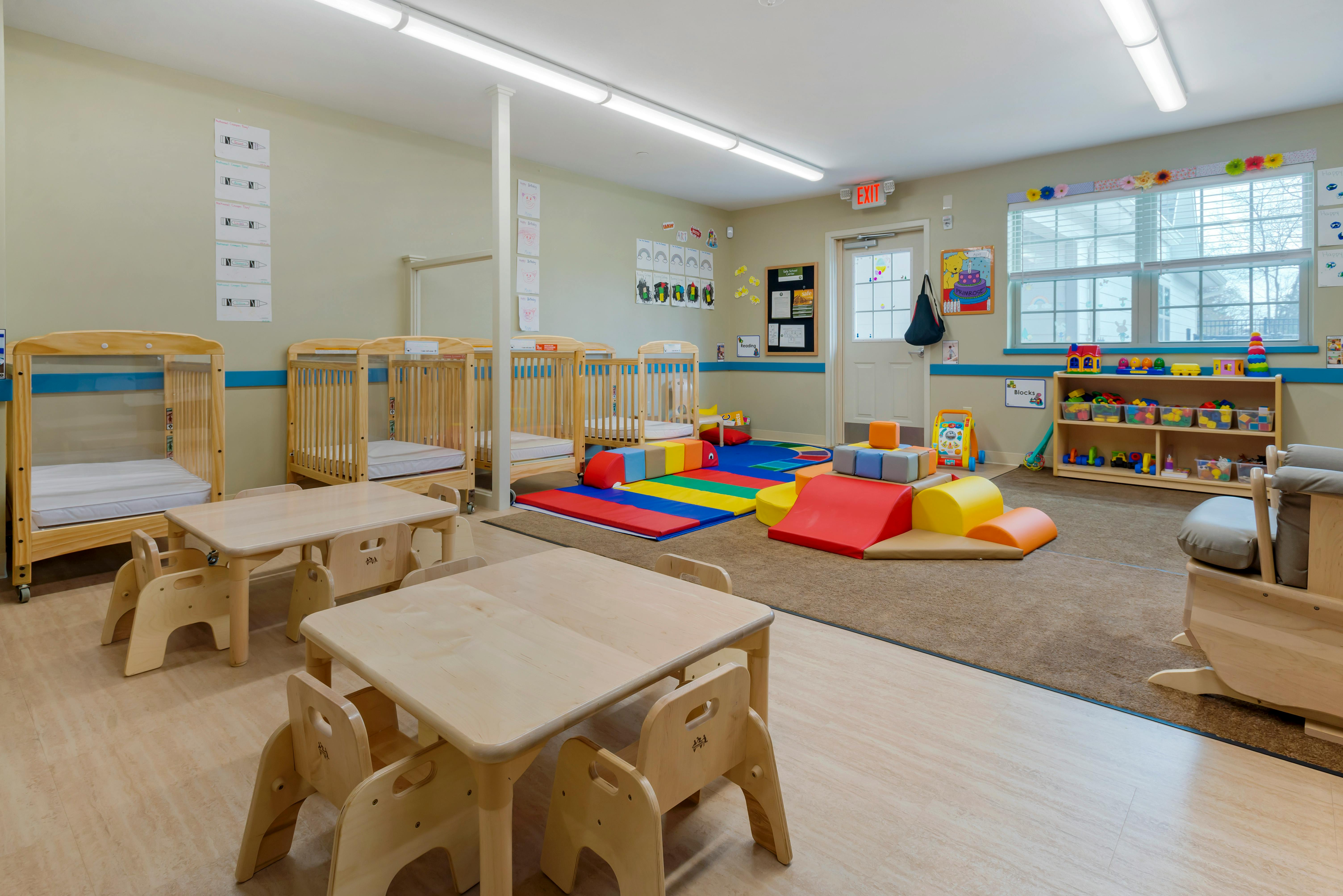 An empty childcare room with cribs, small tables, and colorful play mats