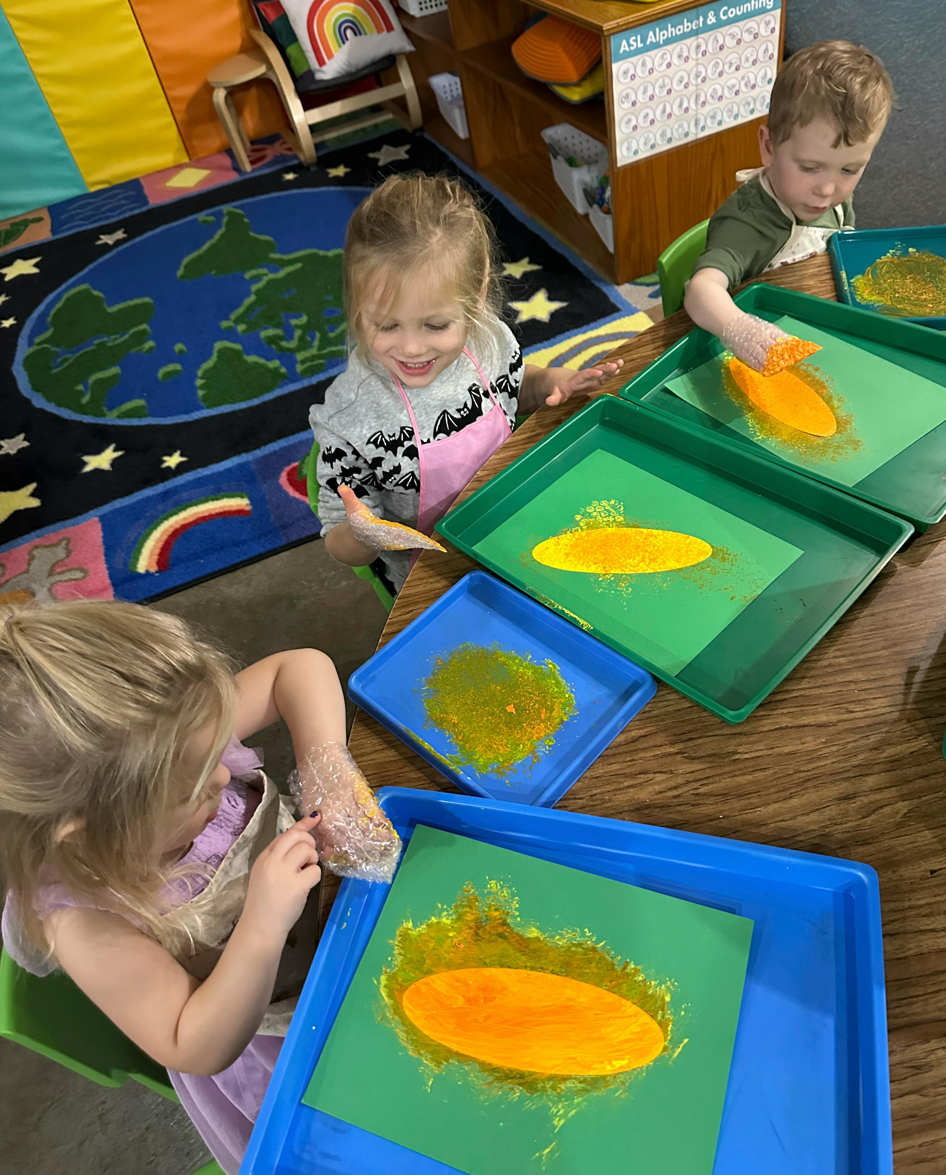 Children wearing plastic gloves painting on trays in a classroom