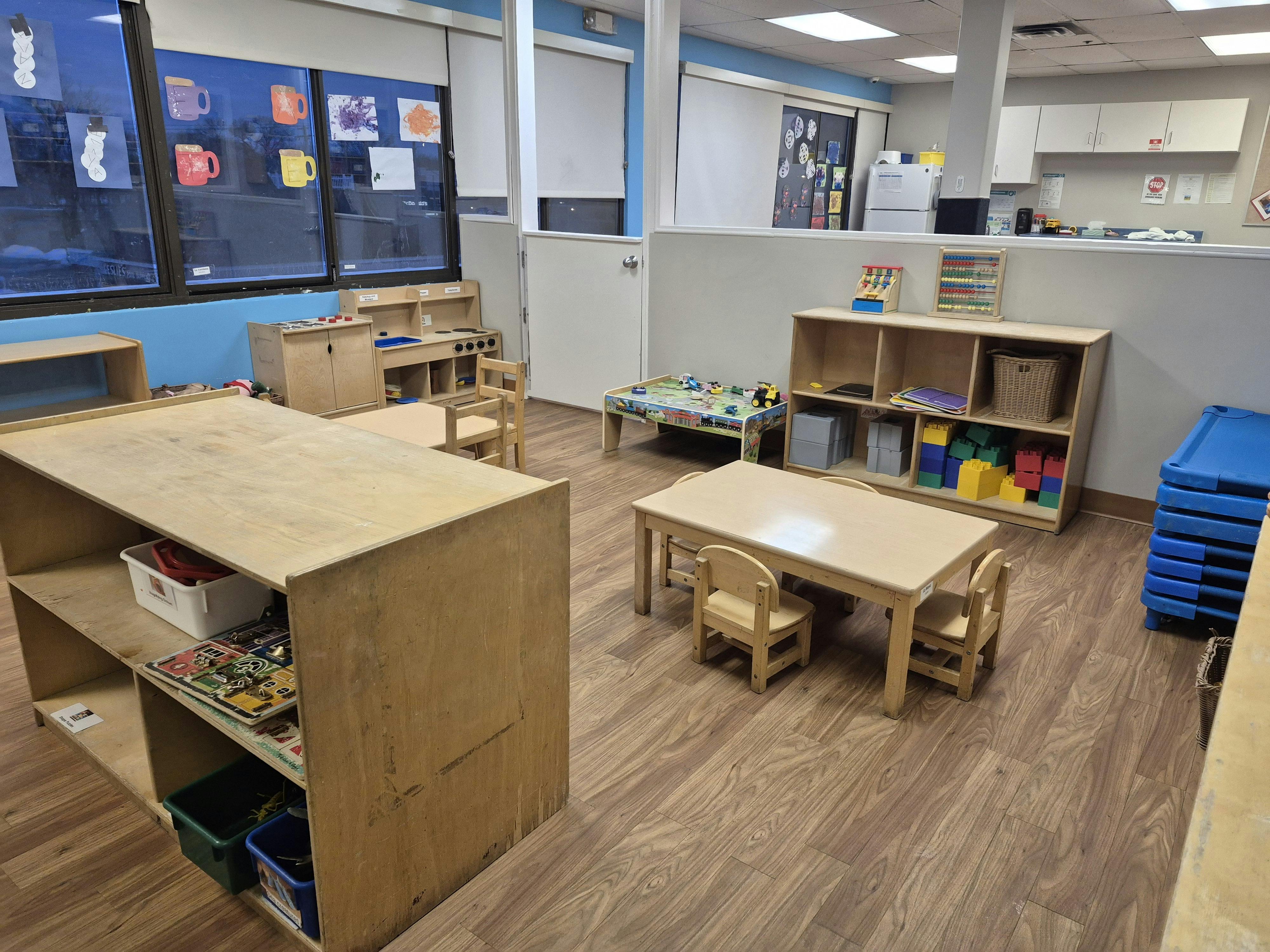 An empty daycare room with small wooden tables, chairs, and play areas