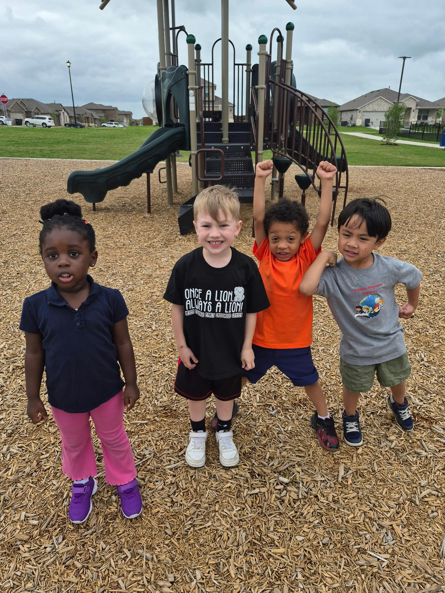 Four children standing on wood chips in front of a playground structure