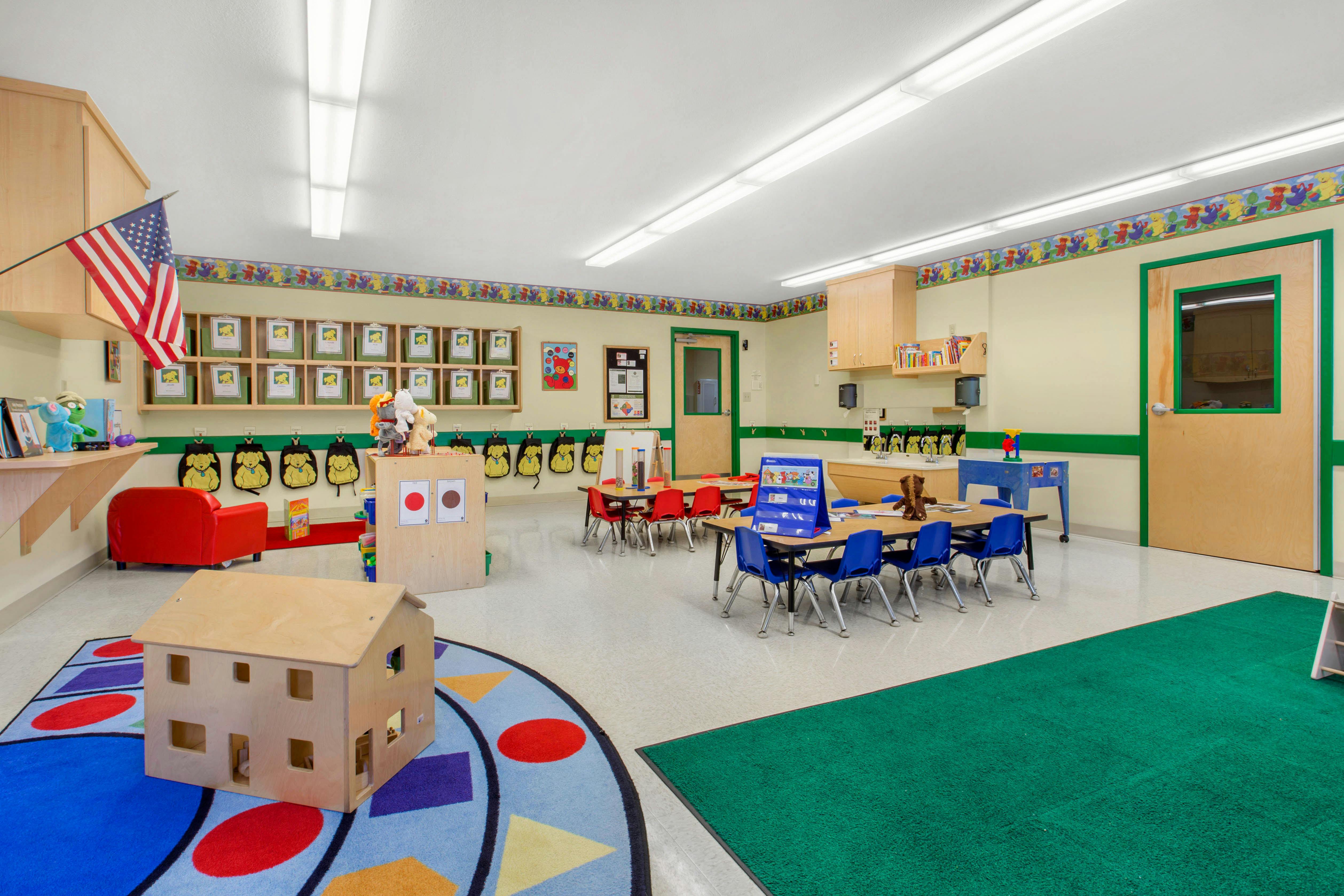 Classroom with small tables and chairs, cubbies, shelves, and a dollhouse on a rug