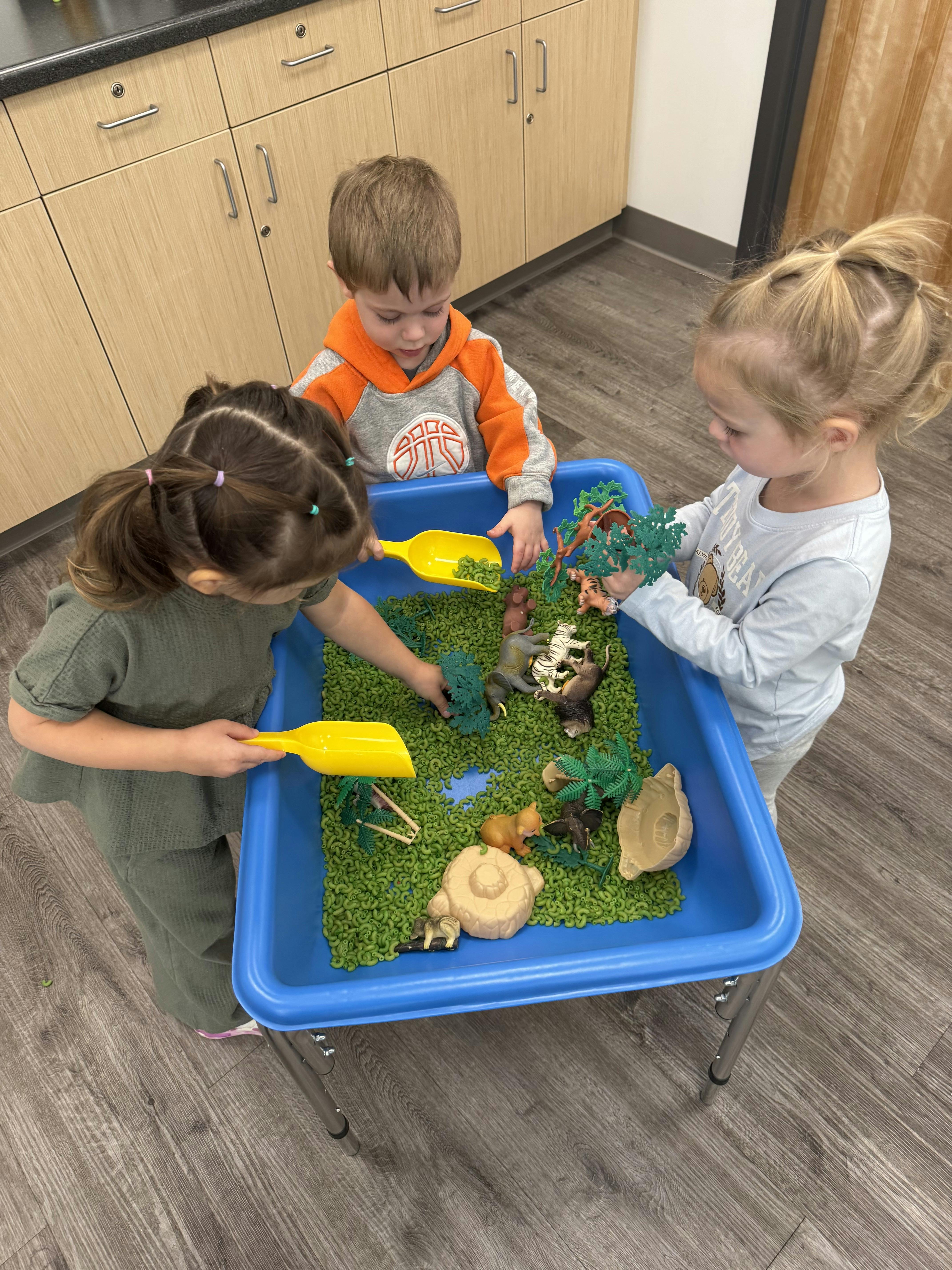 Three children playing with plastic animal figurines in a sensory bin
