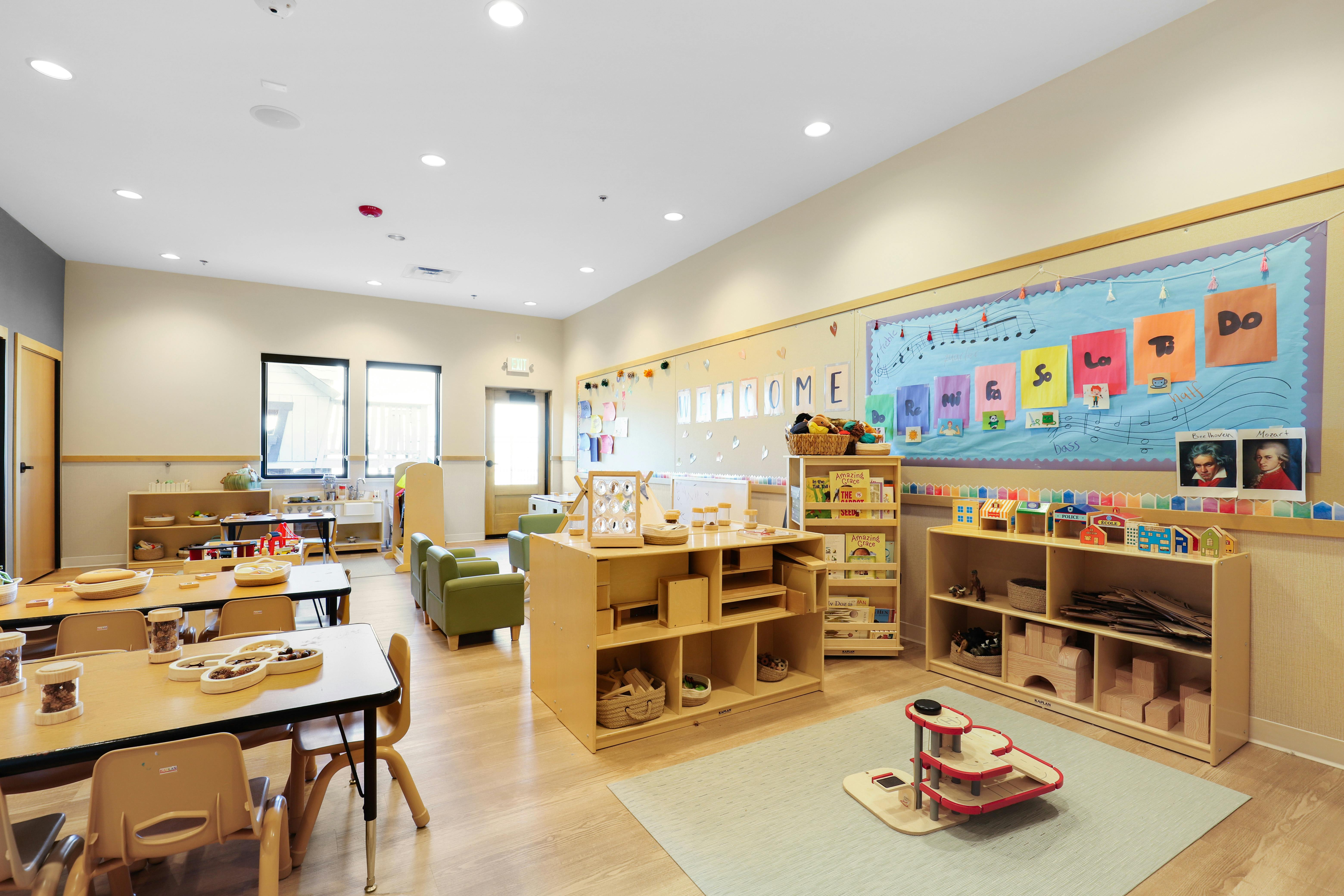 Classroom with small tables and chairs, shelves with toys, and a bulletin board