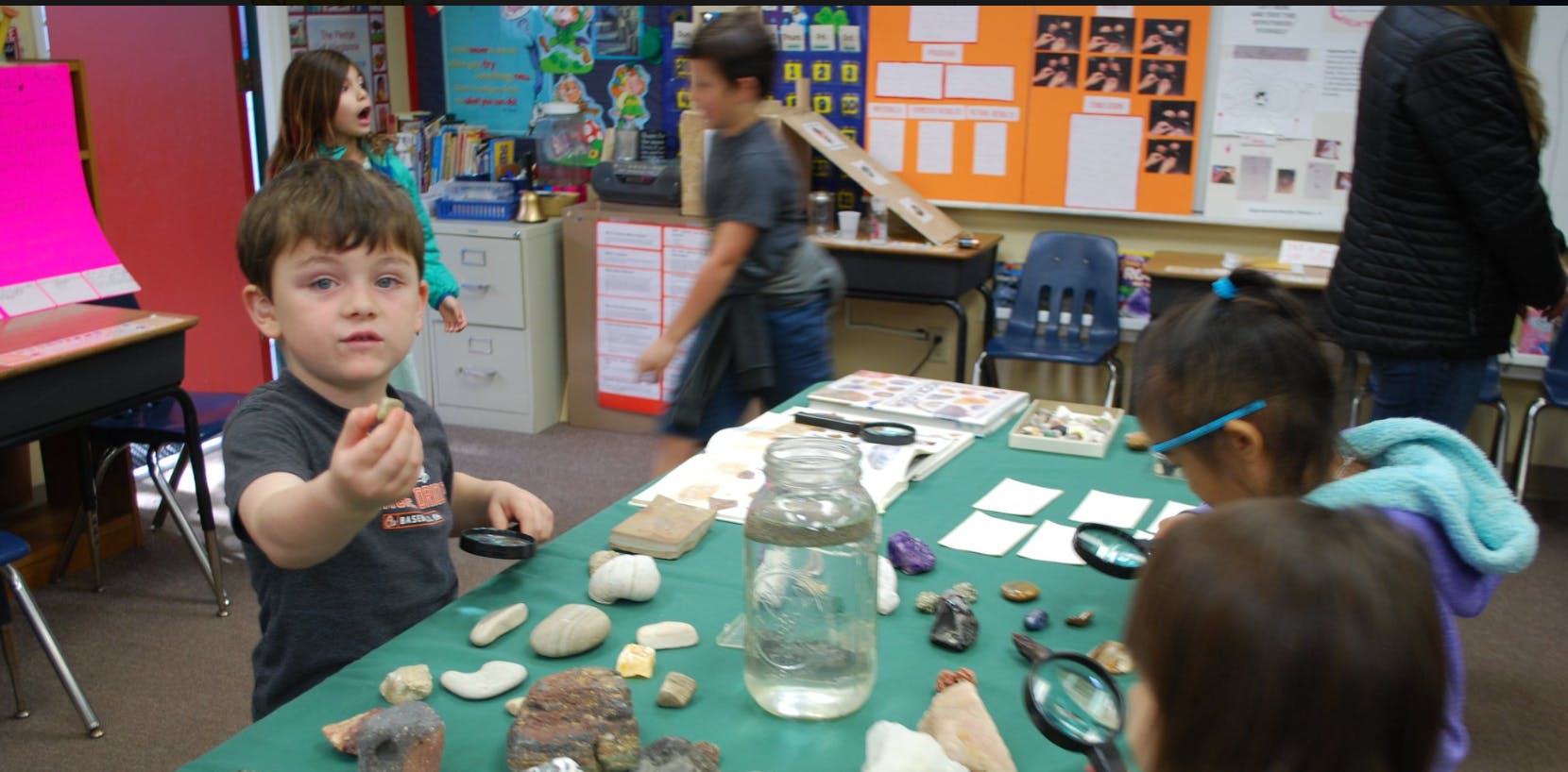 Child holding a small object, other children at a table with rocks and magnifiers