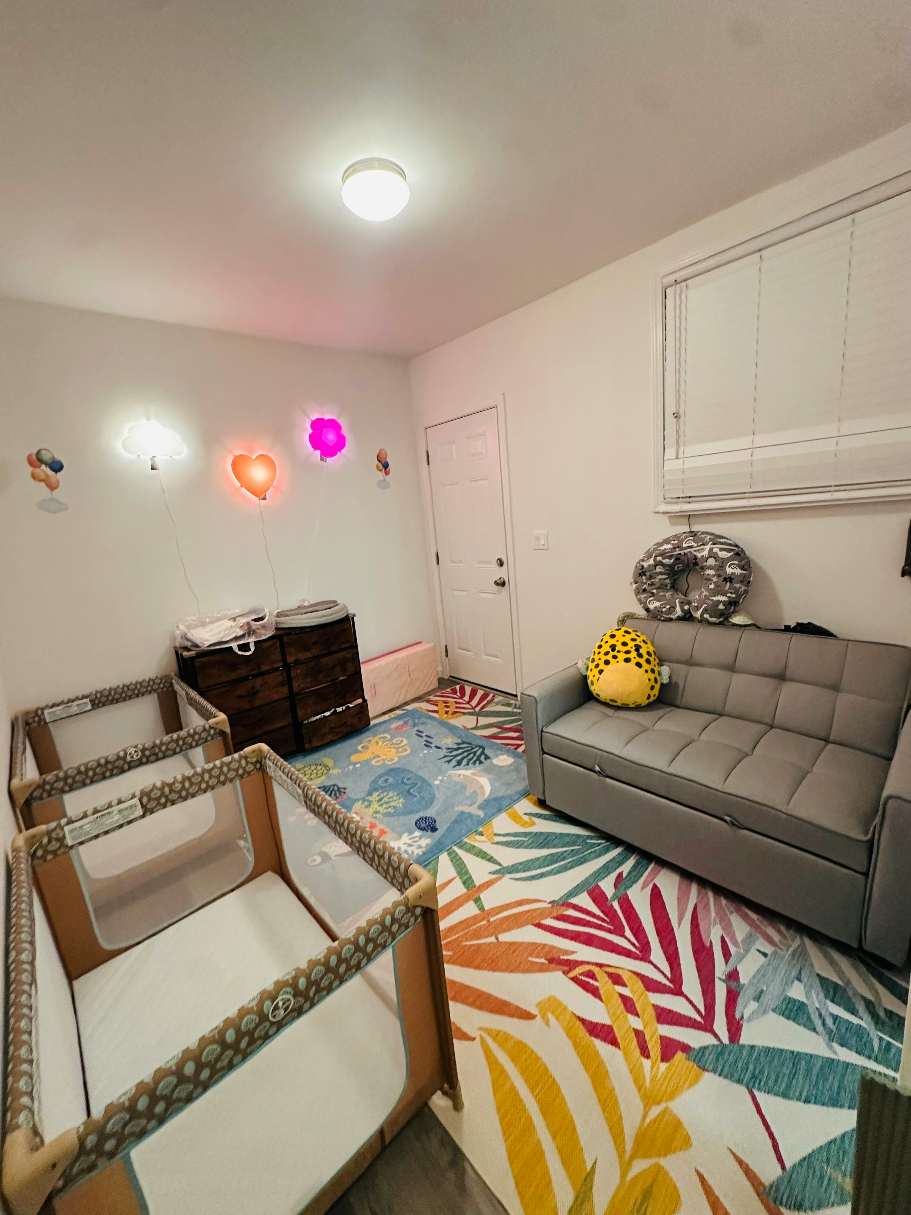 Two cribs, a changing table, and a gray sofa in a room with colorful rugs