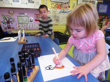 Child drawing at a table in a classroom with another child in the background