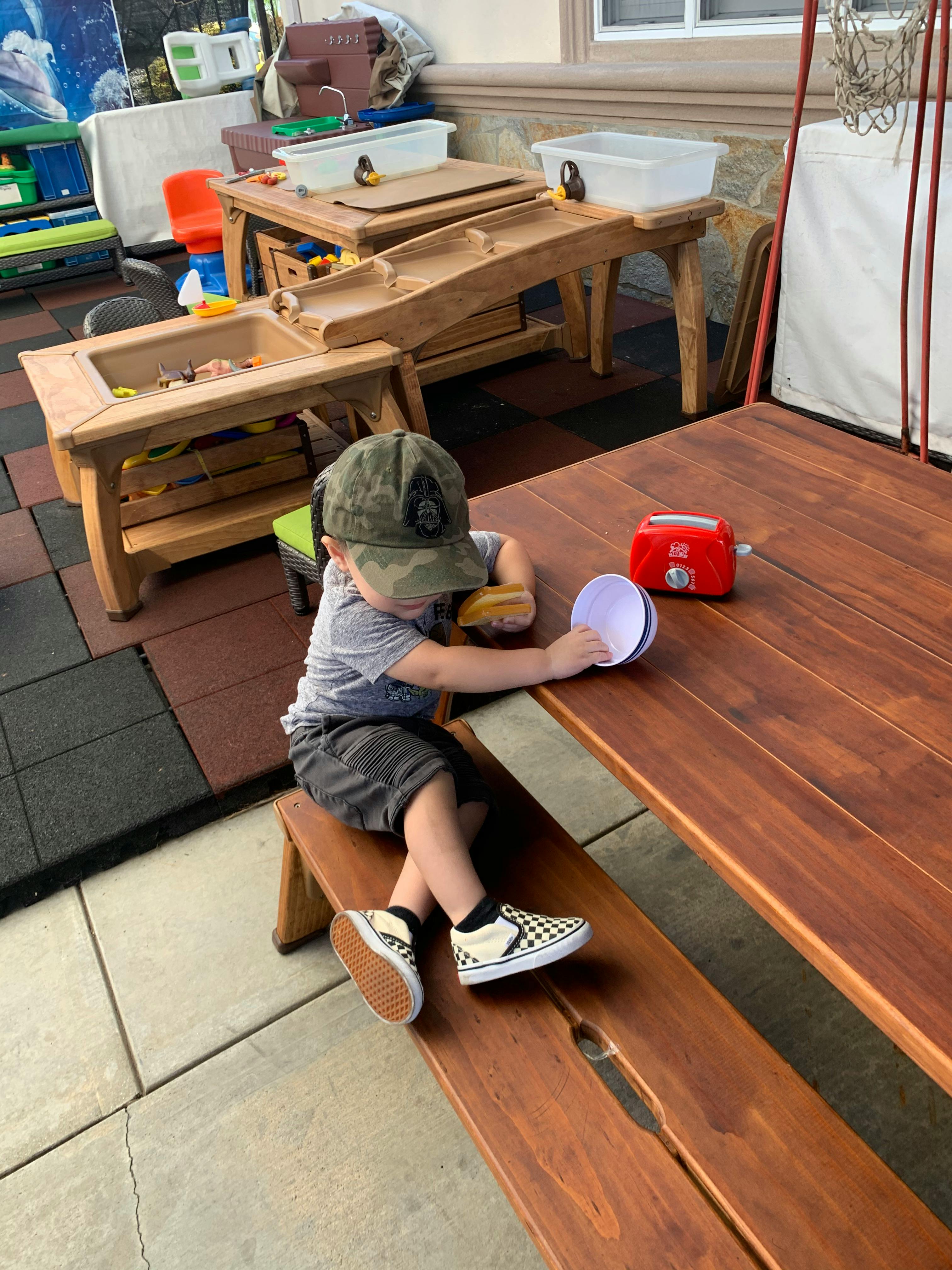 Child in a camouflage hat playing with a toy toaster and bowl on a wooden picnic bench