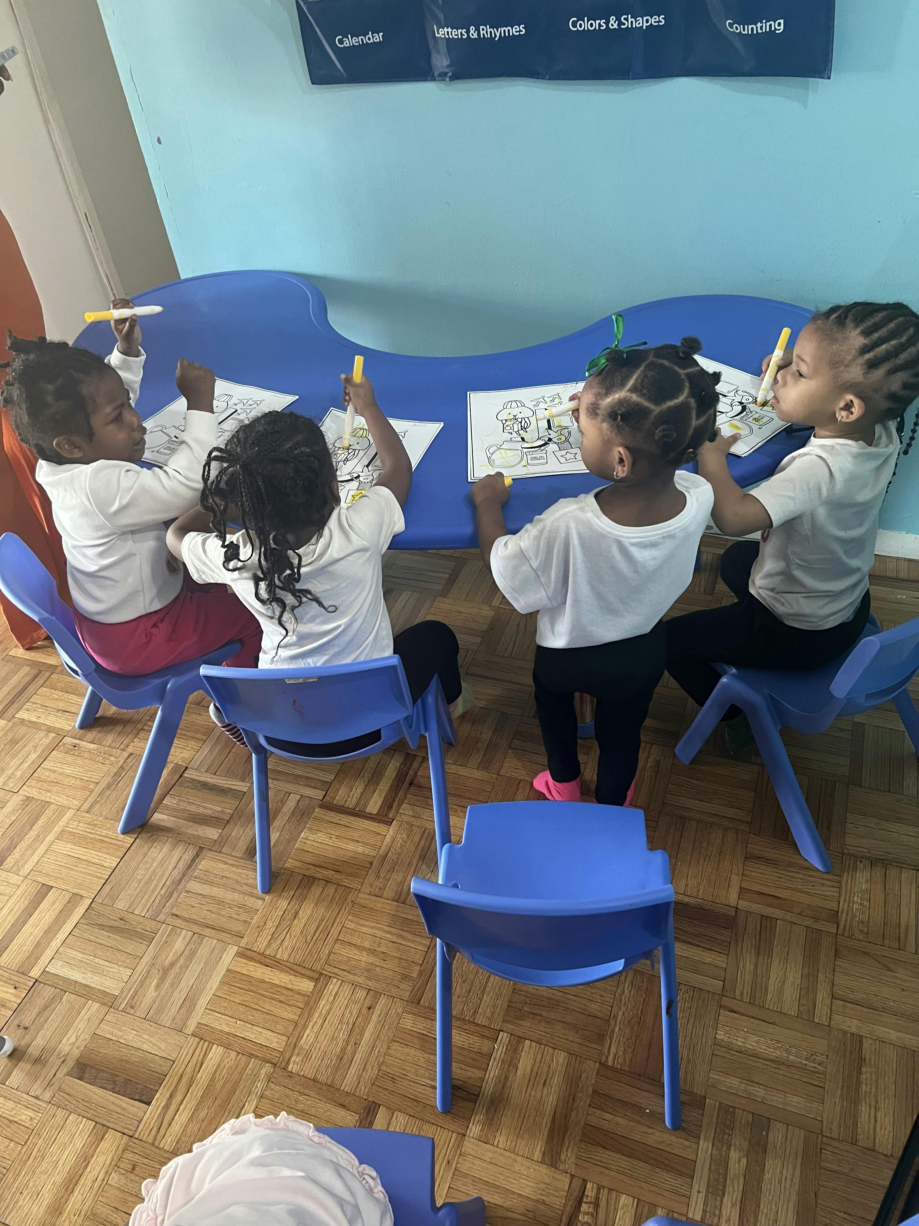 Four children coloring on papers at a blue table in a classroom