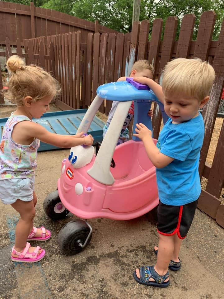 Three children playing with a pink toy car outdoors