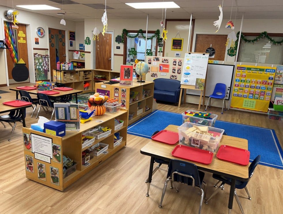 Classroom with small tables, chairs, shelving units with supplies, and learning posters
