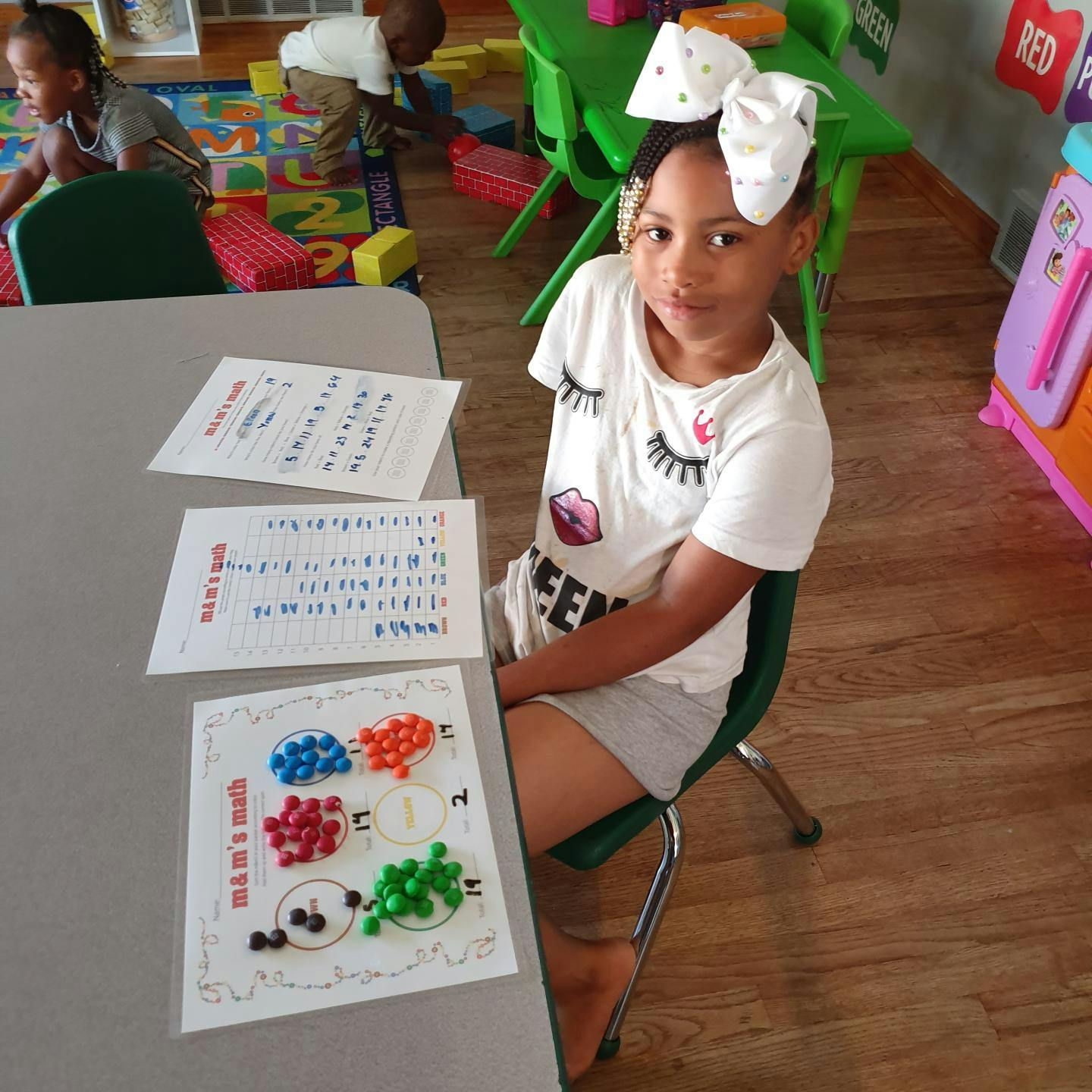 Child sitting at a table with papers and colorful objects in a classroom