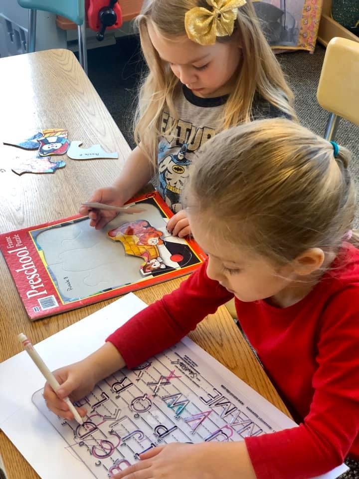 Two children at a table, one coloring a worksheet, the other playing with a puzzle
