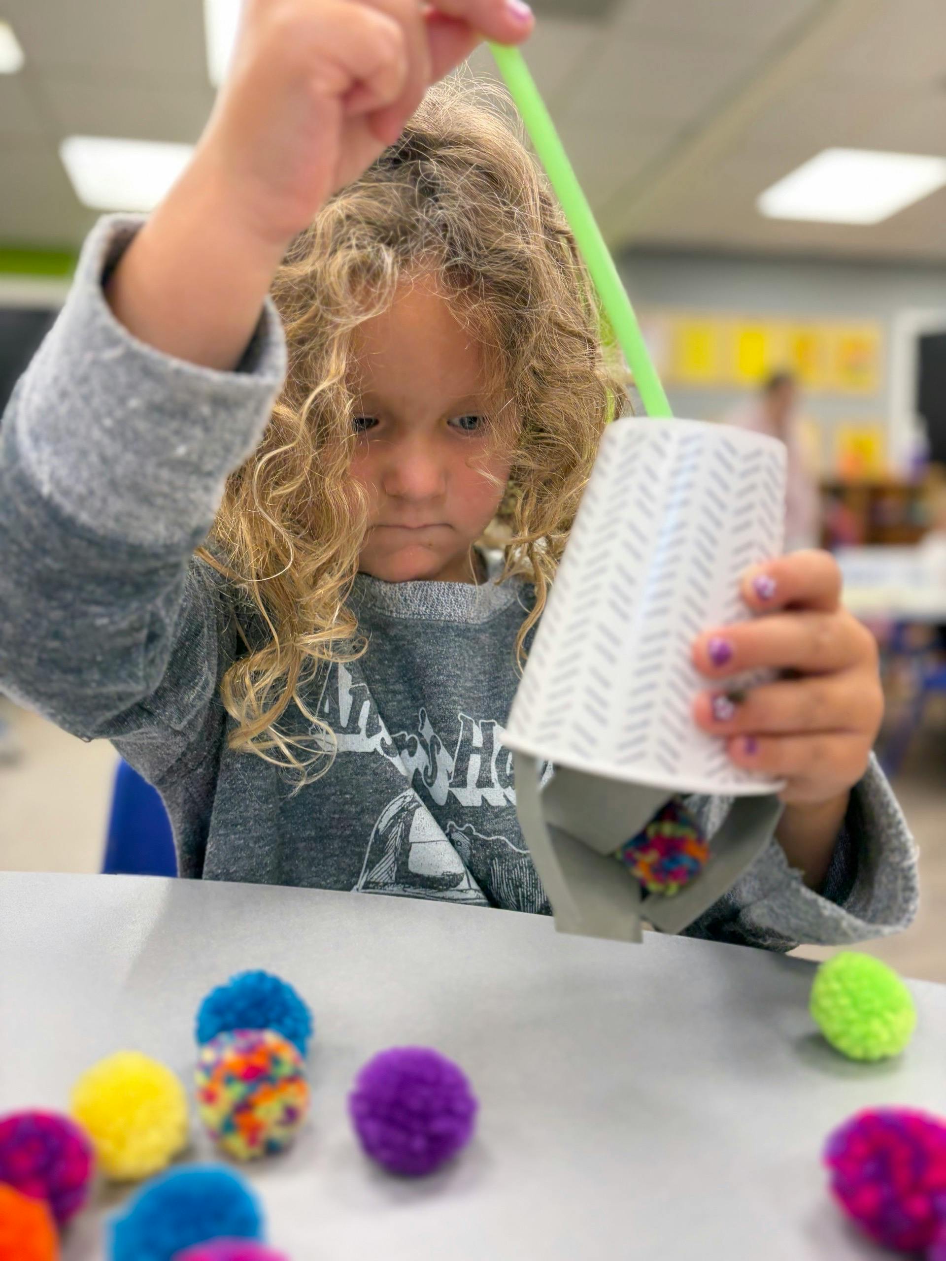 Child using a straw and paper cup to pick up small pom-poms