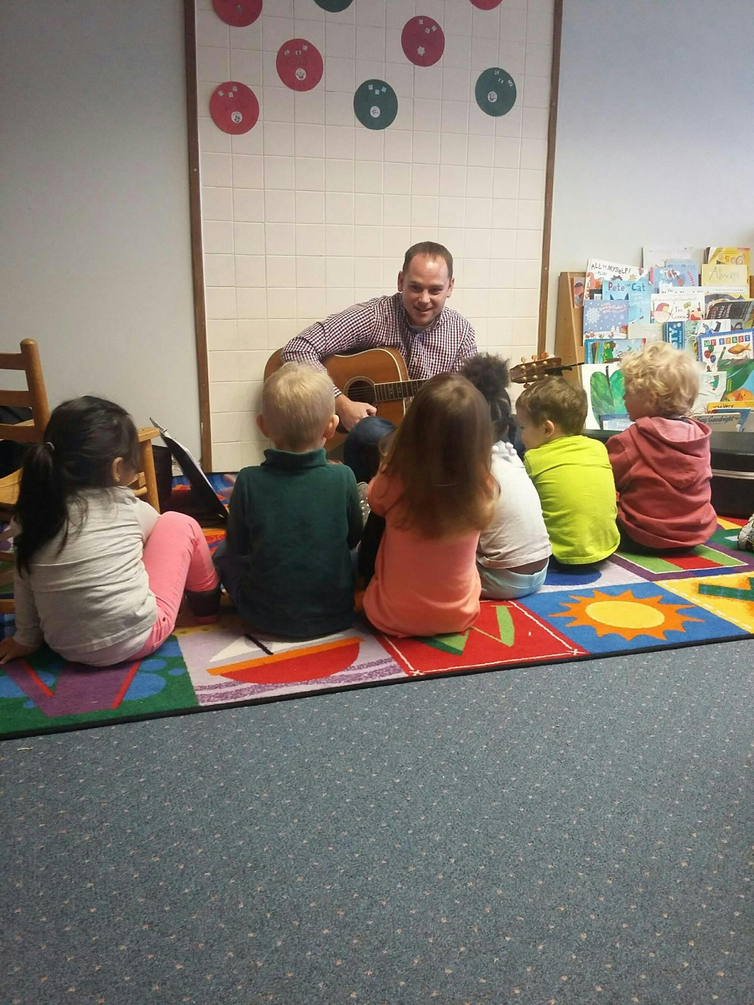 Adult playing guitar for a group of children sitting on a colorful rug