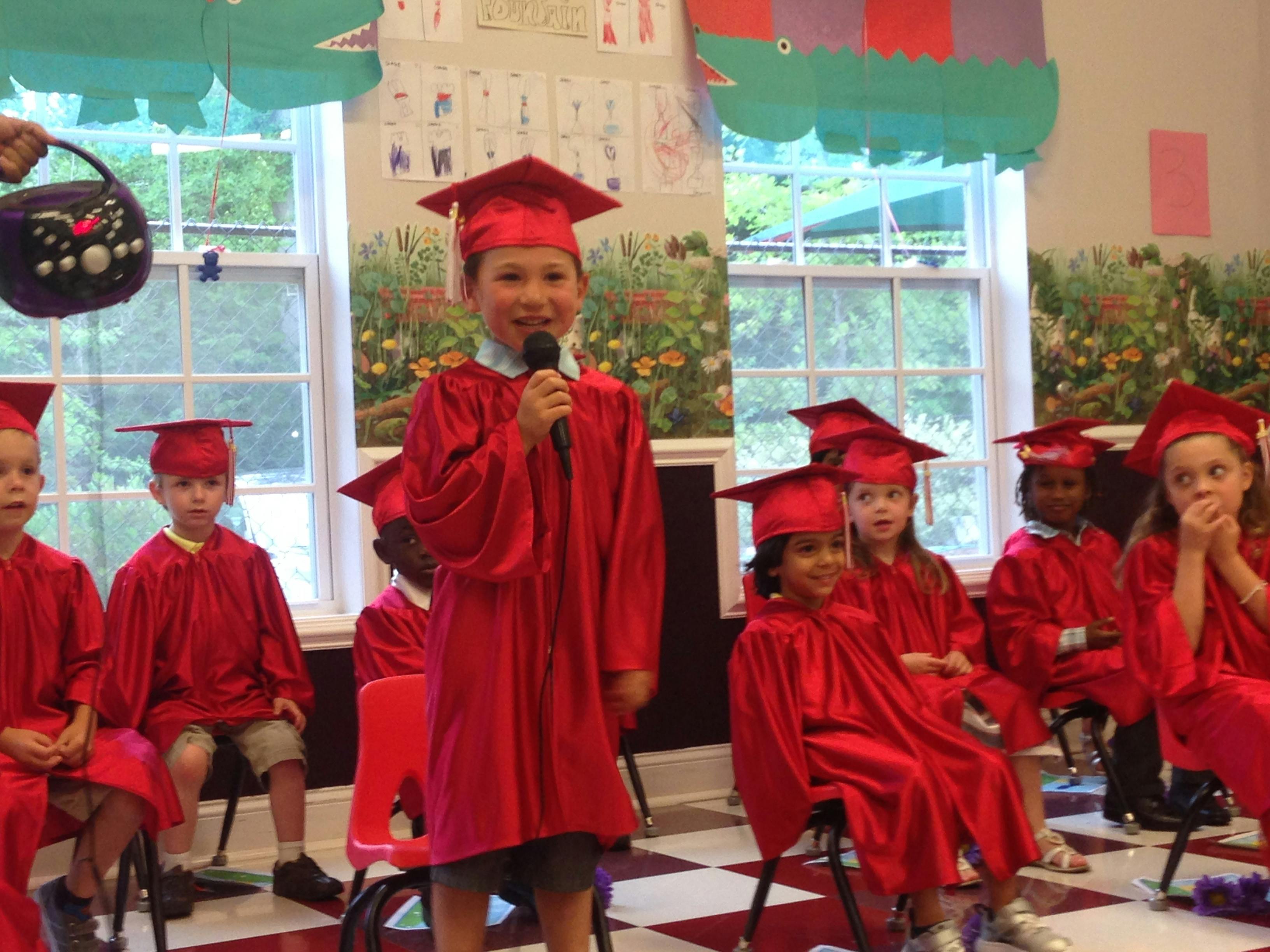 Child in red graduation gown speaking into a microphone, other children seated