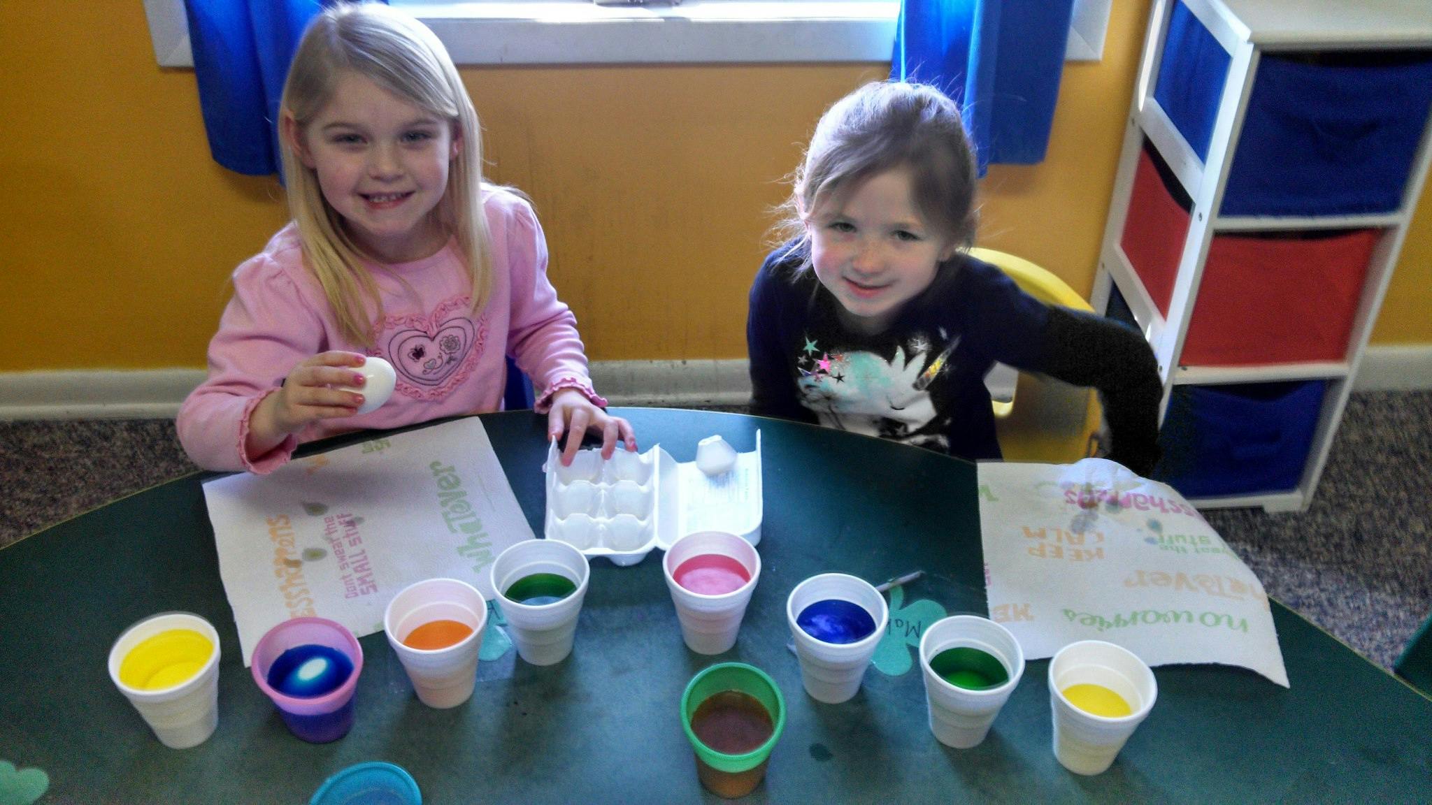 Two children at a table with cups of colored liquid and an egg carton