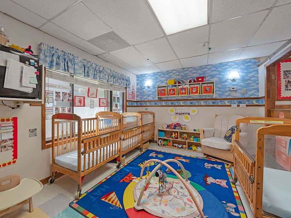 A childcare classroom with multiple cribs, a playmat on a blue rug, and shelves