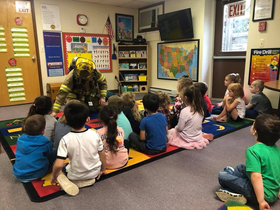 Person in firefighter gear kneeling in front of children in a classroom