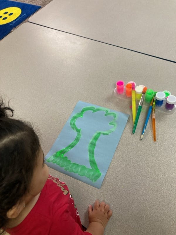 Child painting a tree outline on blue paper with paint and brushes nearby