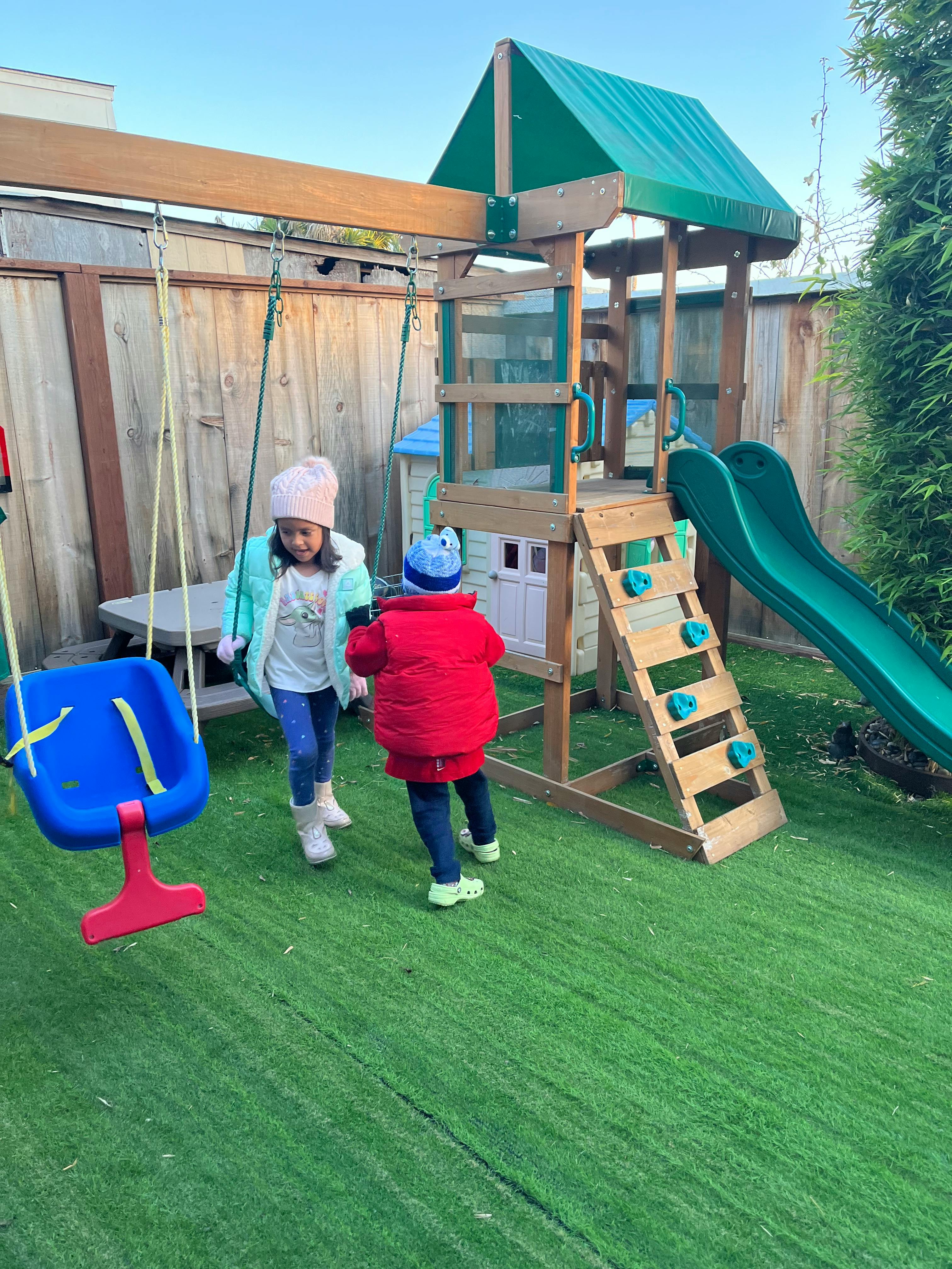 Two children playing on a backyard playset with a slide and swings