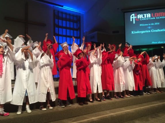 Children in red and white graduation gowns with hands raised