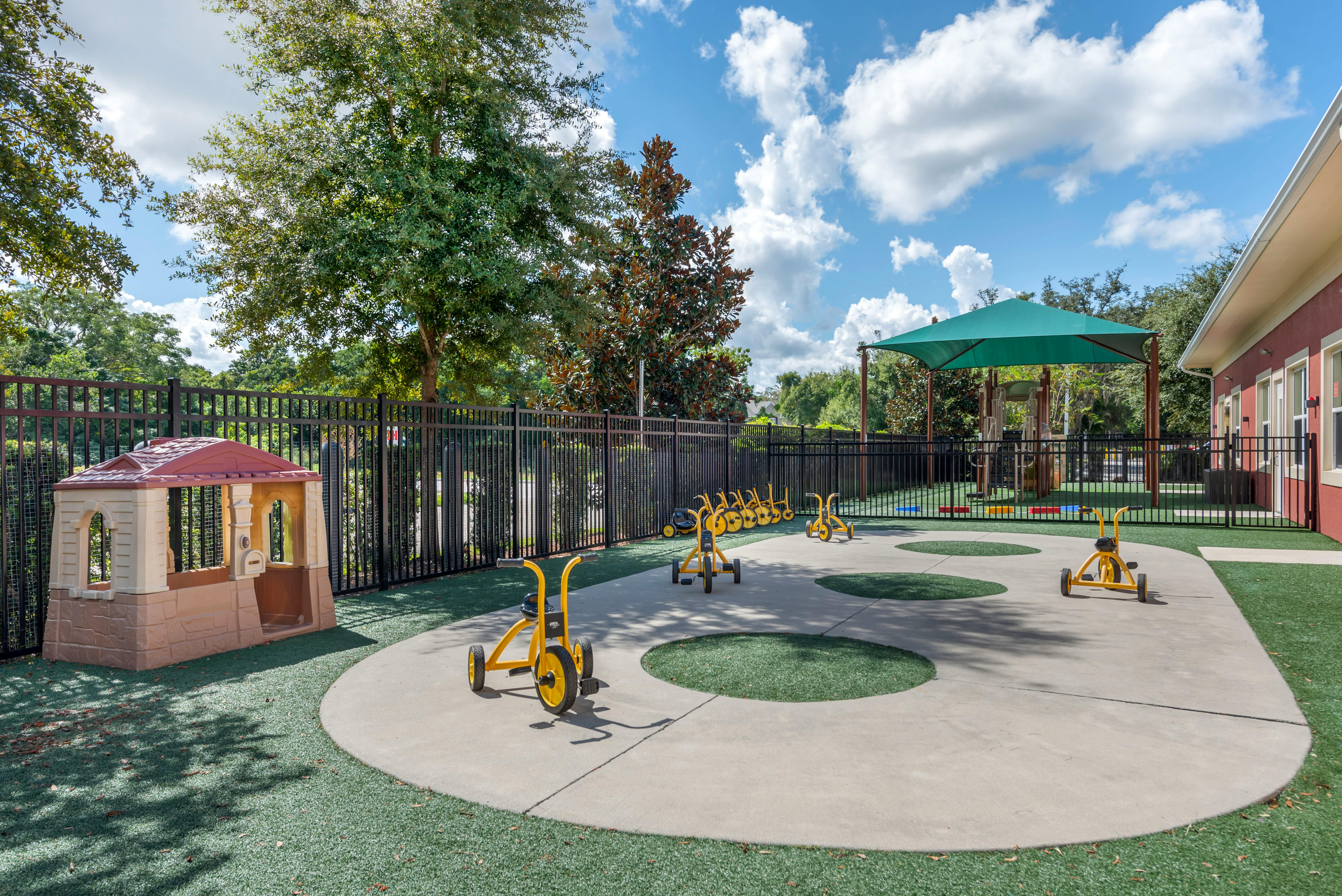 Outdoor playground with a playhouse, tricycles, and a shaded play structure