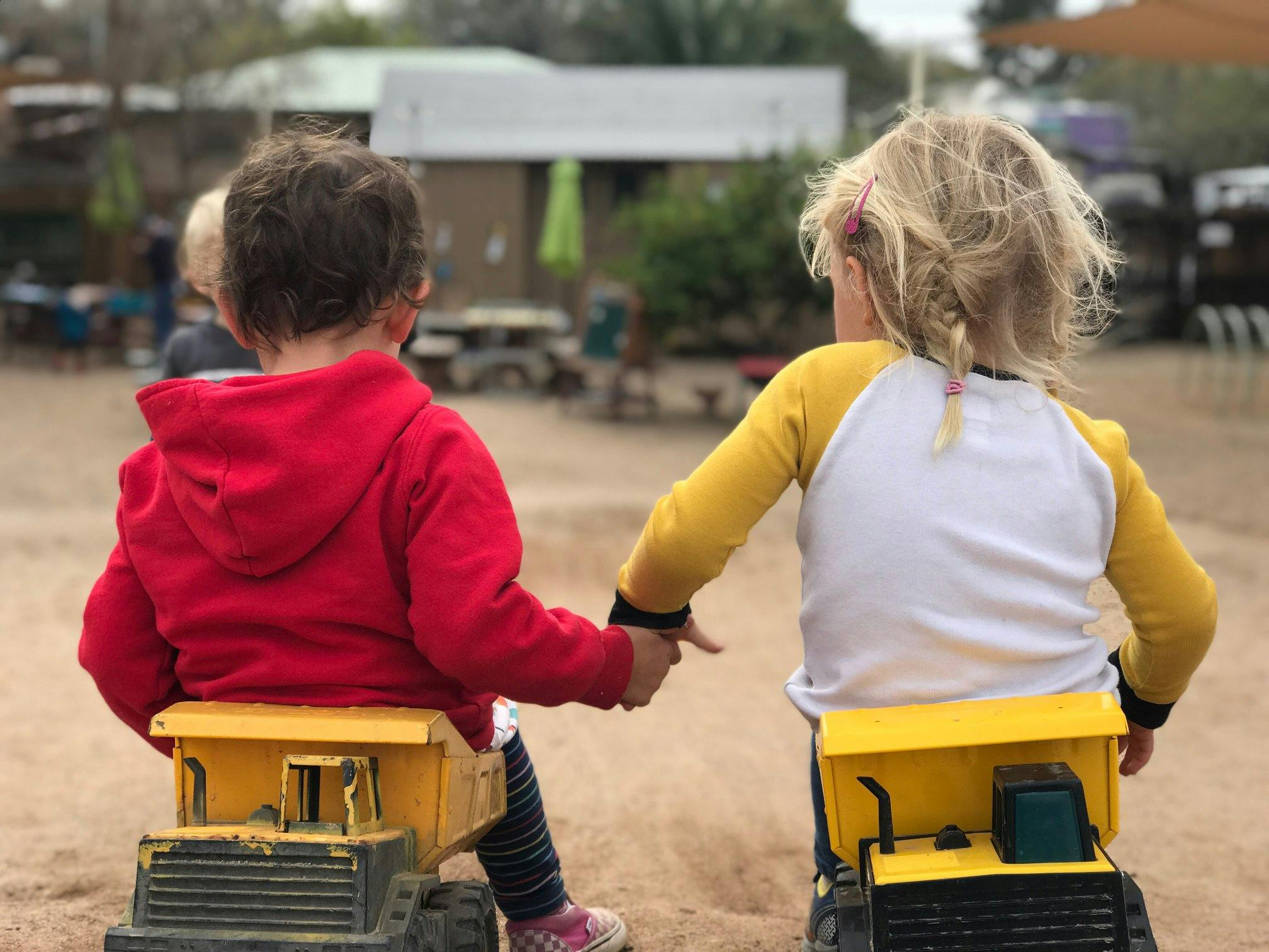 Two children holding hands while sitting on toy trucks