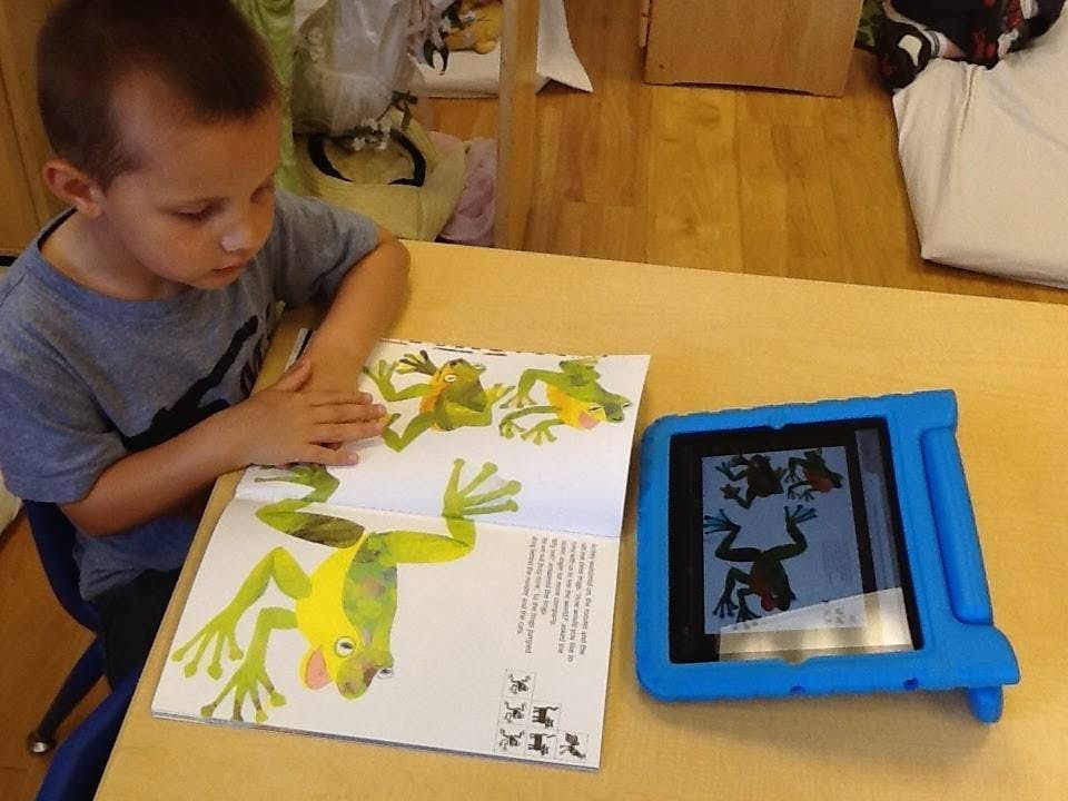 Child reading a book with frog illustrations next to a tablet
