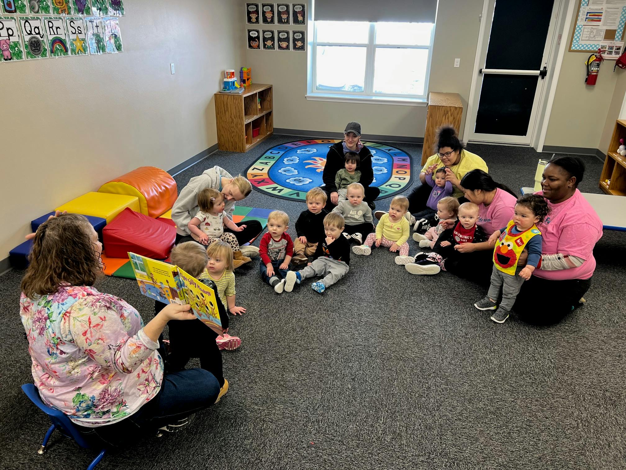 Adults and children sitting on the floor in a classroom, one adult reading a book