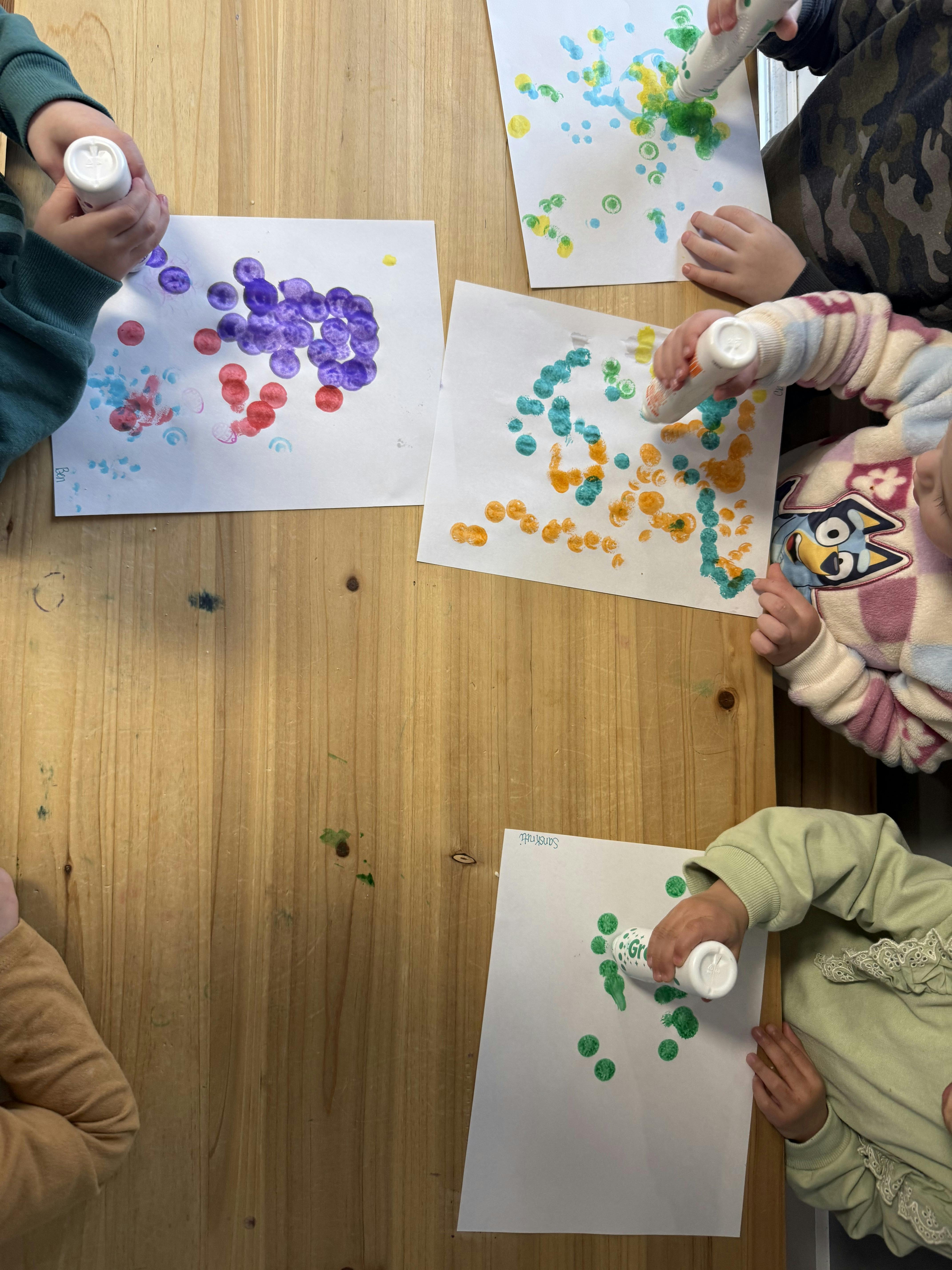 Multiple children's hands using dot markers to paint on white paper at a wooden table