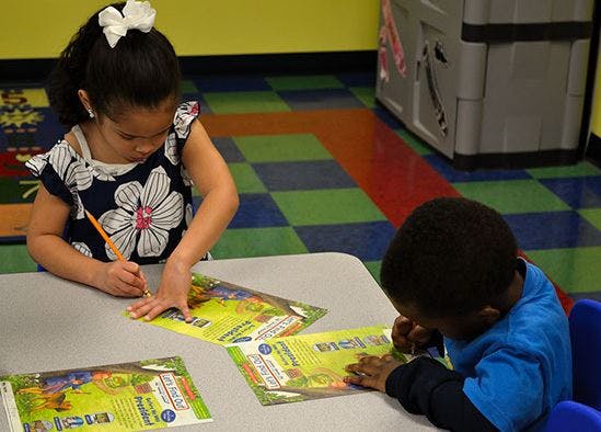 Two children at a table, one writing and one looking at a book