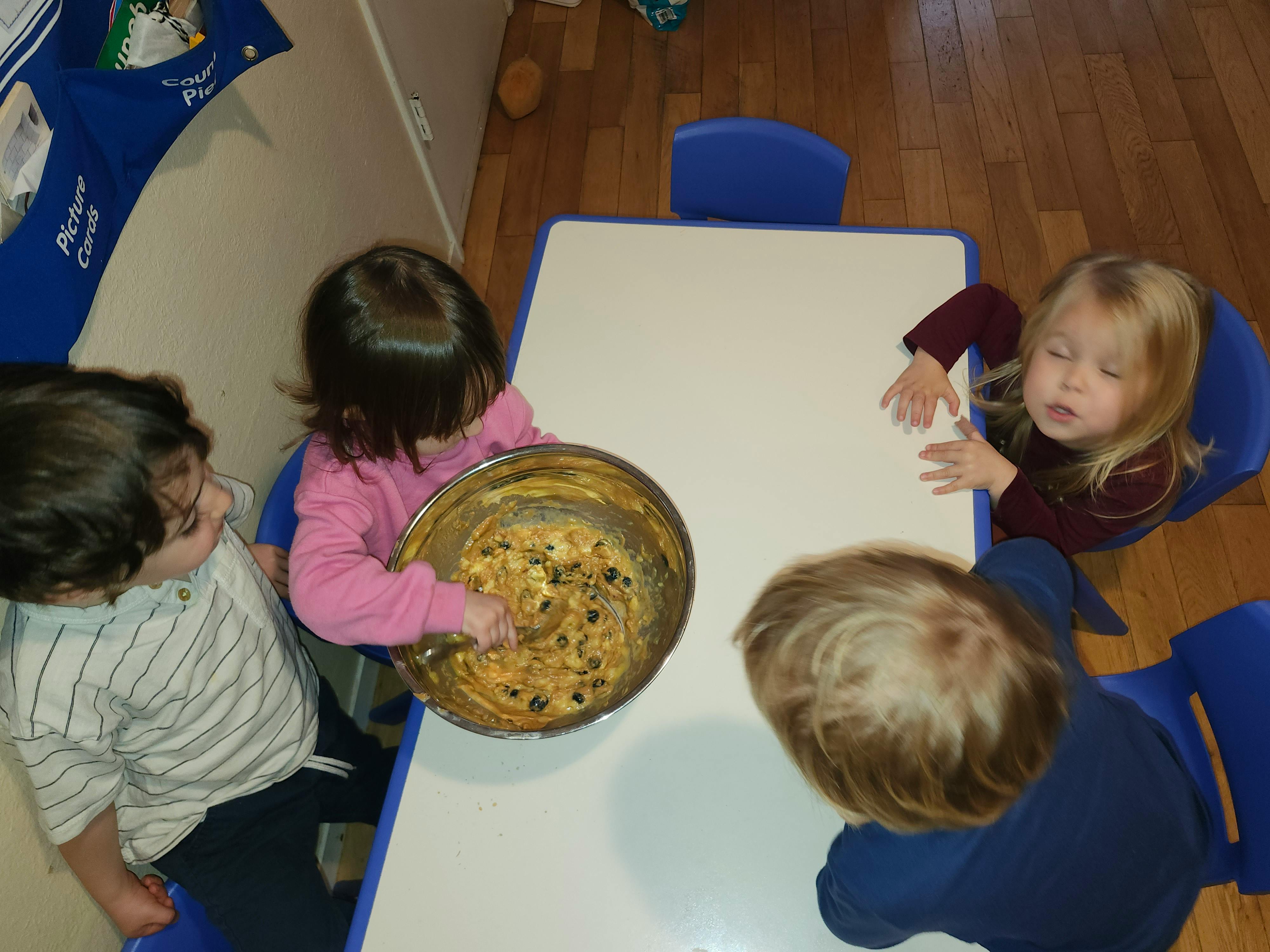 Three children sitting at a table with one child stirring batter in a bowl