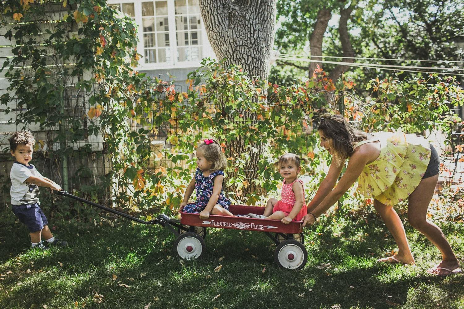 A person and three children playing with a red wagon in a yard