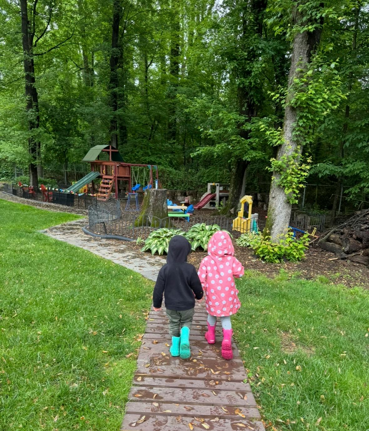 Two children in rain boots walking on a path towards a playground