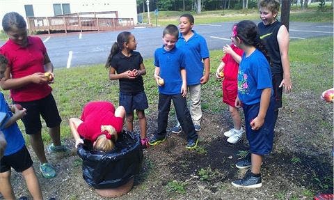 Children bobbing for apples in a black barrel outdoors