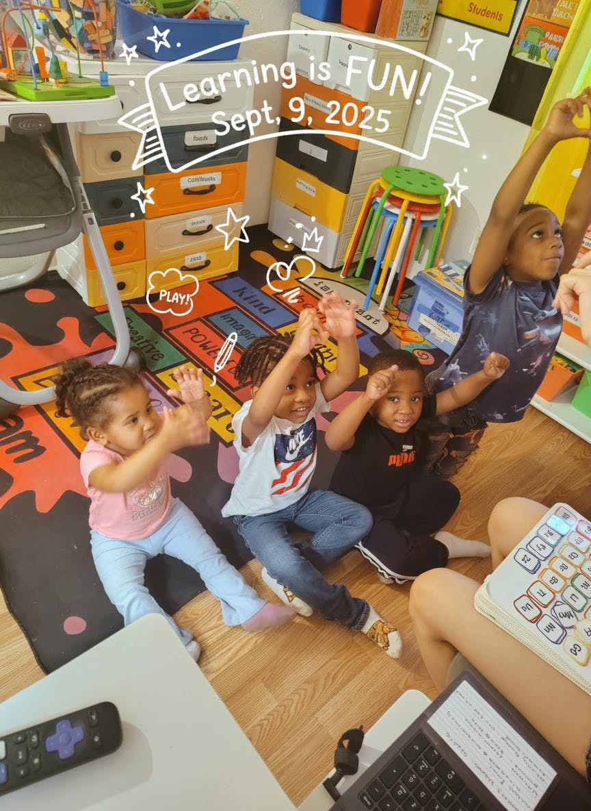 Four children sitting on the floor with arms raised, in a classroom