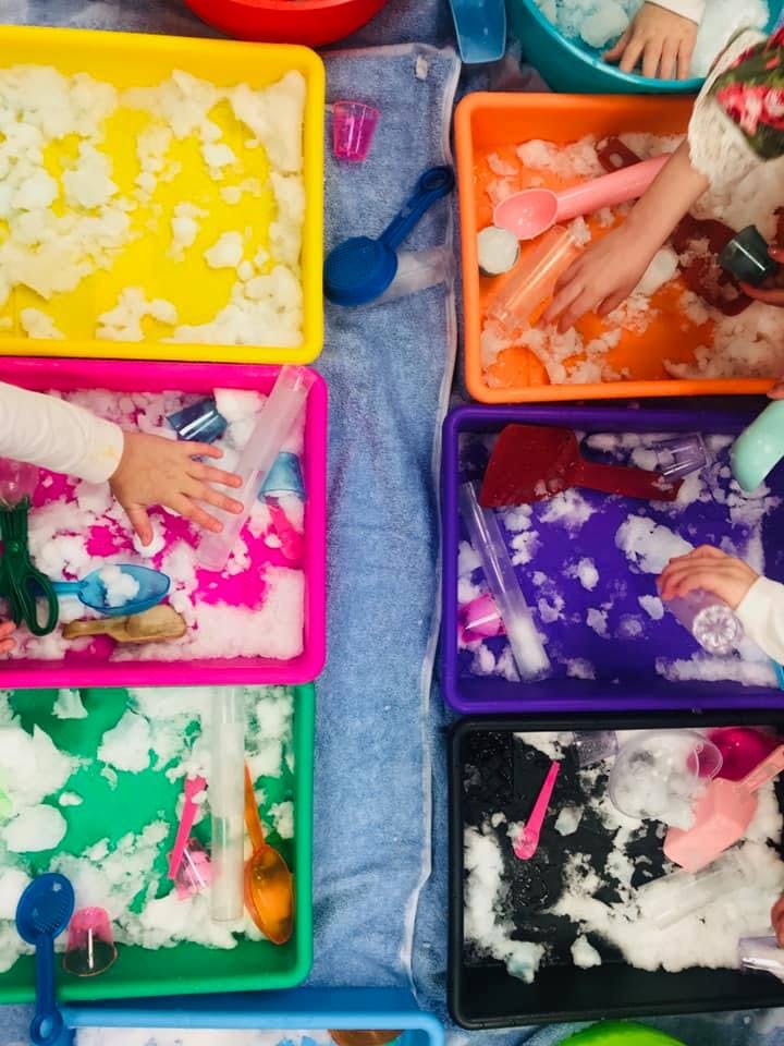 Multiple colorful bins with snow and various objects, with children's hands playing