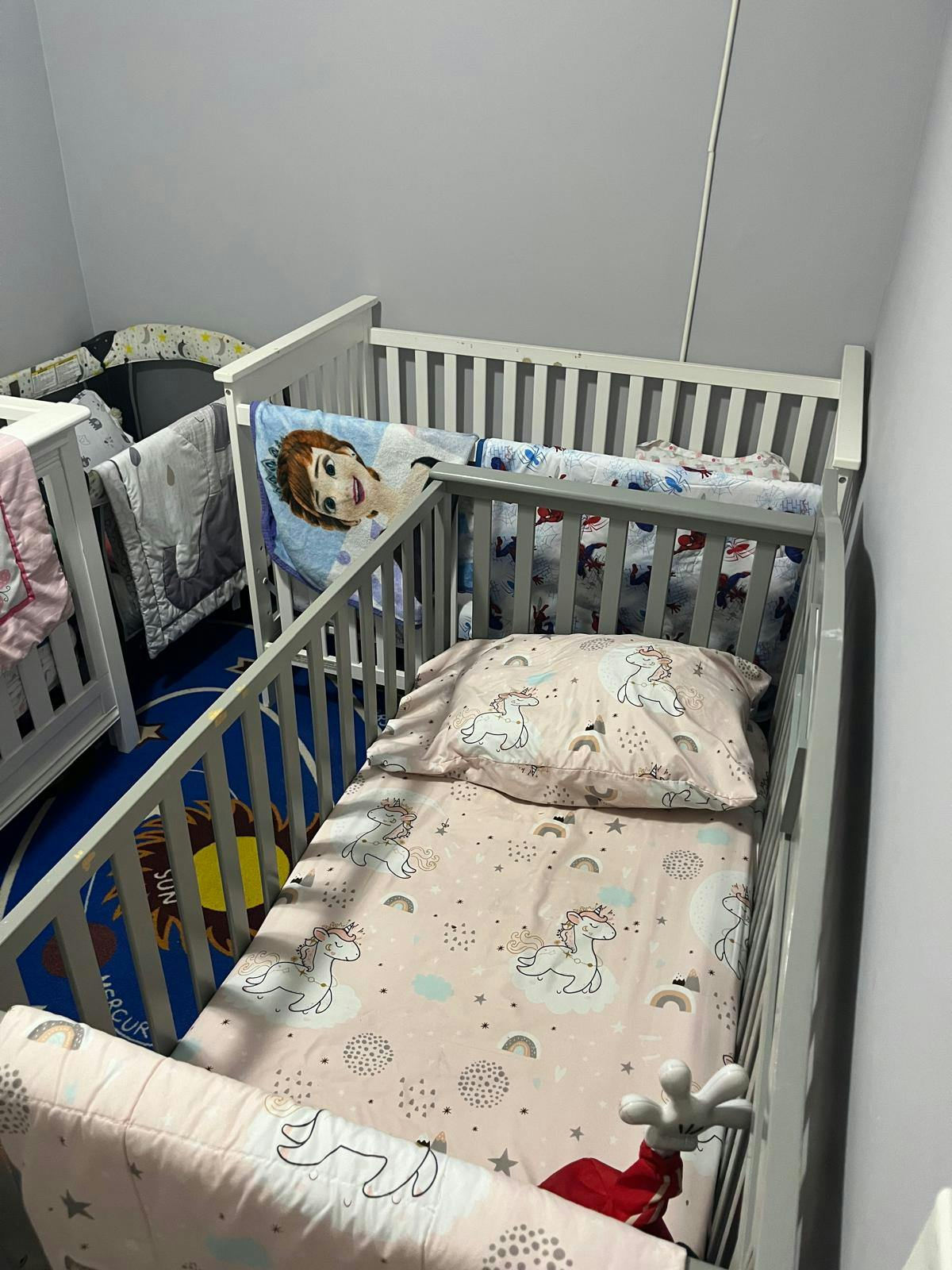 Two cribs with bedding, one with a unicorn pattern and the other with a cartoon character