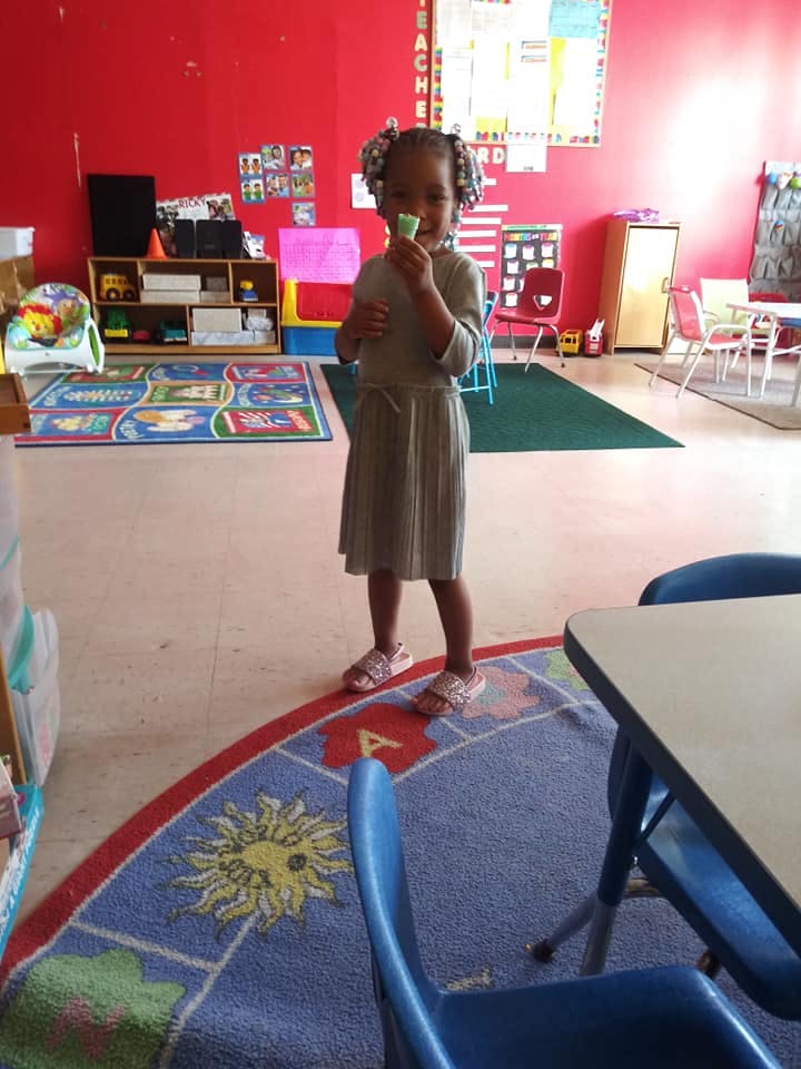 Child standing in a classroom holding a small object to their mouth