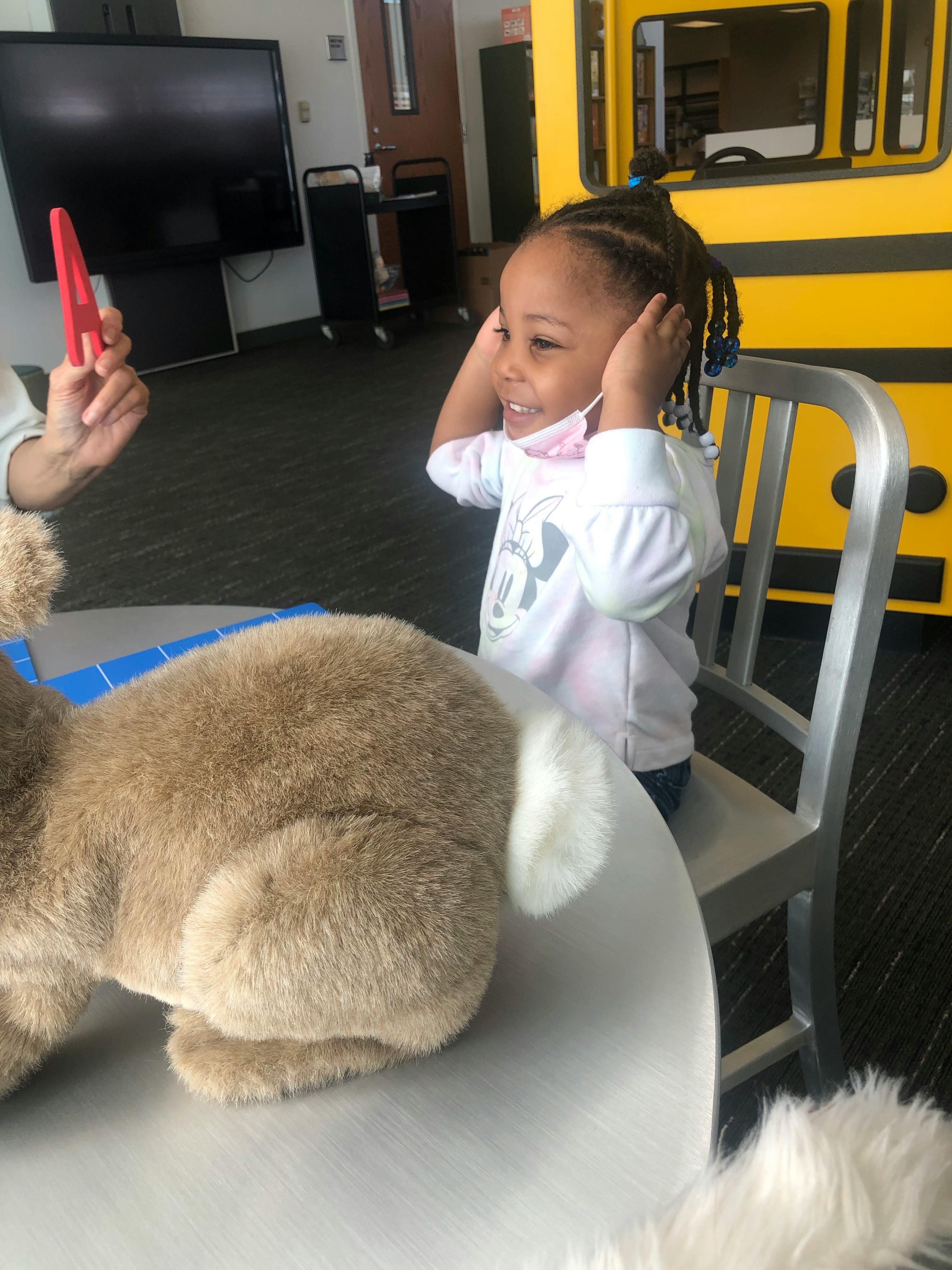 Child sitting at a table with a stuffed animal, looking at a person holding a red object