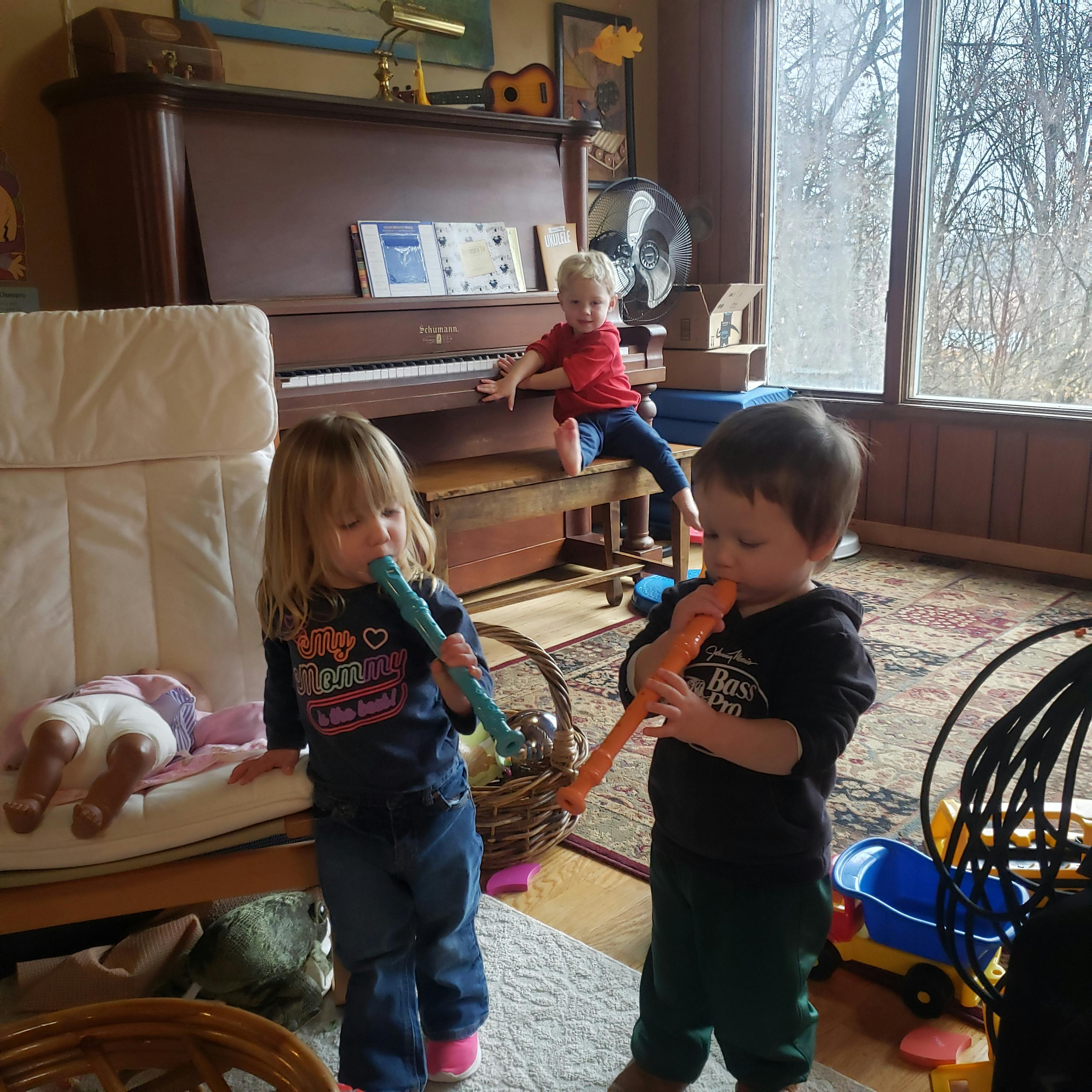Two children playing toy flutes, another child sitting on a piano bench