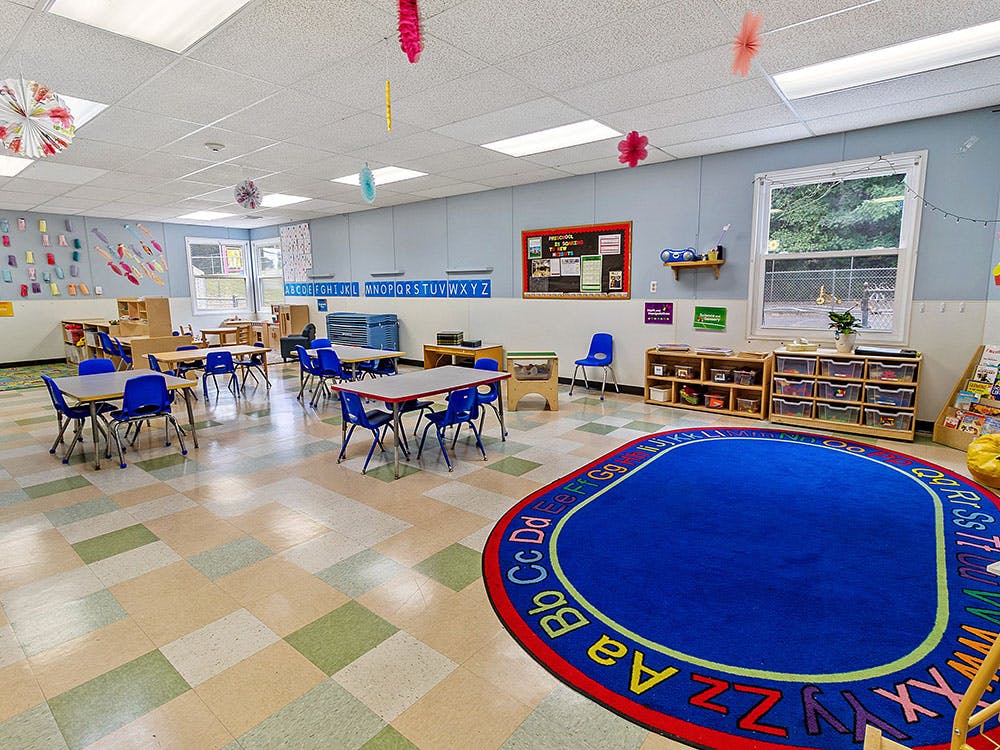Empty classroom with small tables and chairs, and a blue alphabet rug