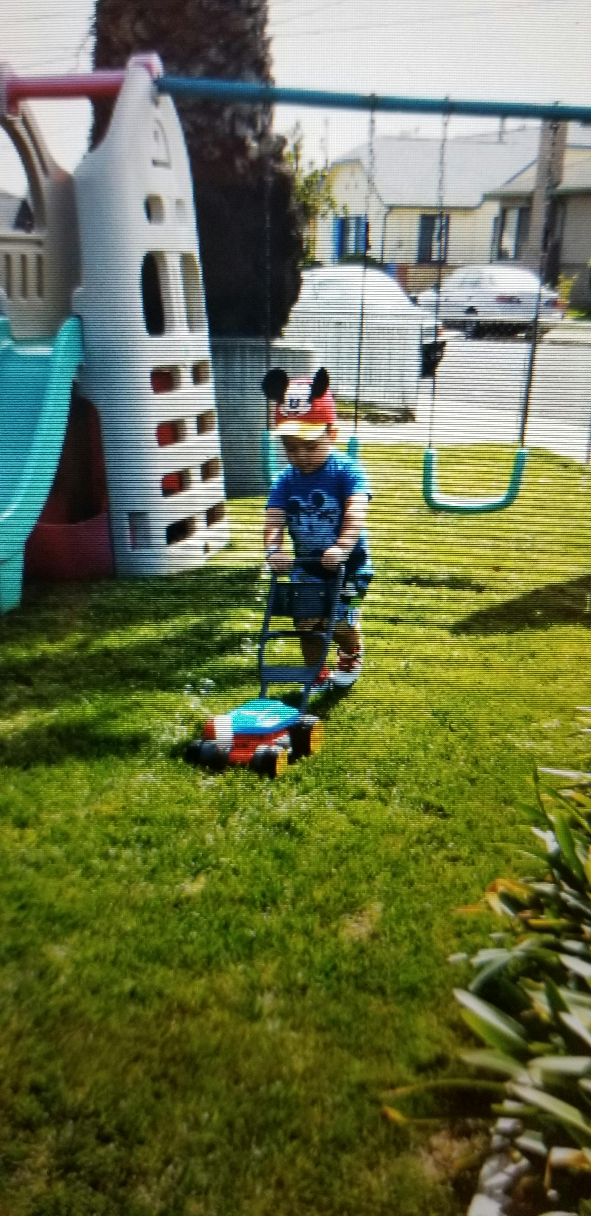Child in a Mickey Mouse hat pushing a toy lawnmower on grass