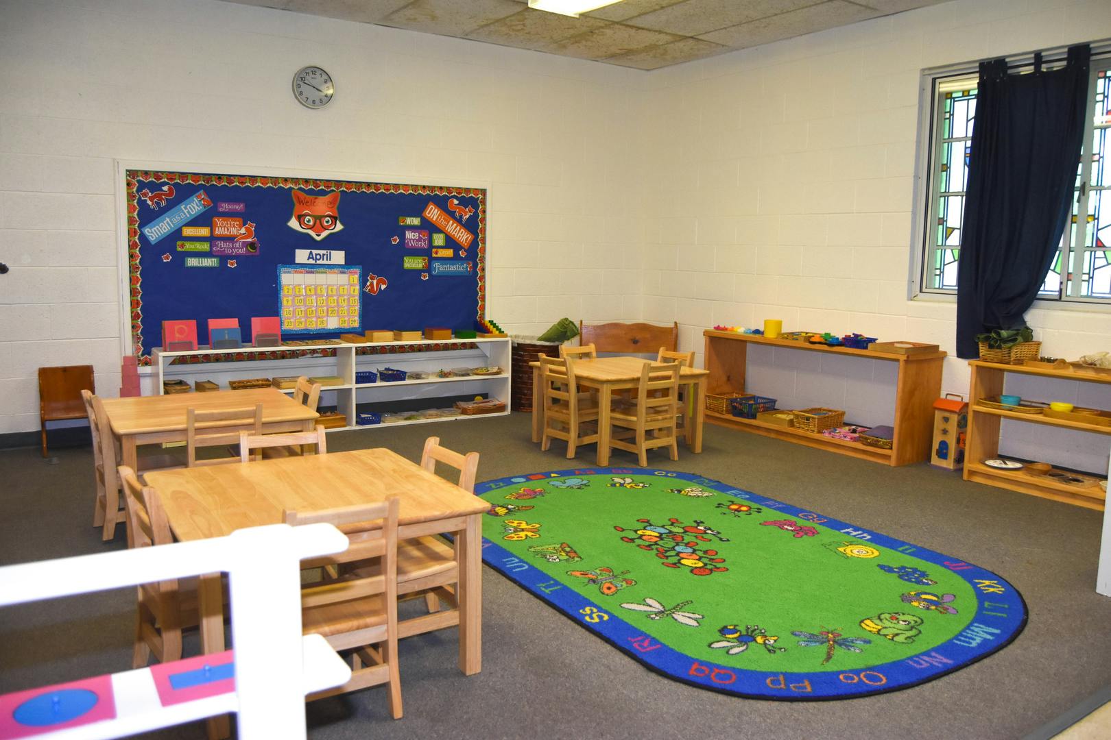 Classroom with small wooden tables and chairs, a green rug, and shelves